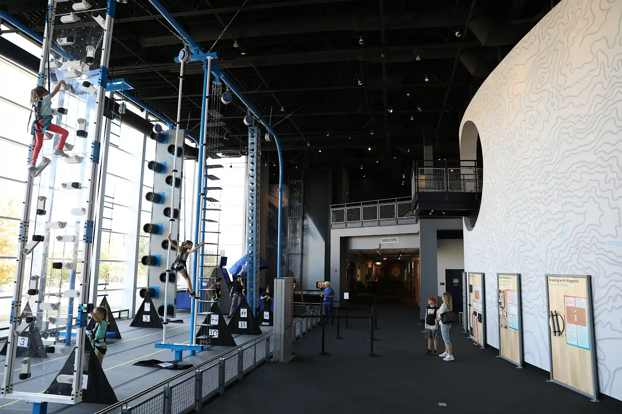 Indoor climbing area with children climbing on various wall structures next to a white wall with contour lines and educational displays.