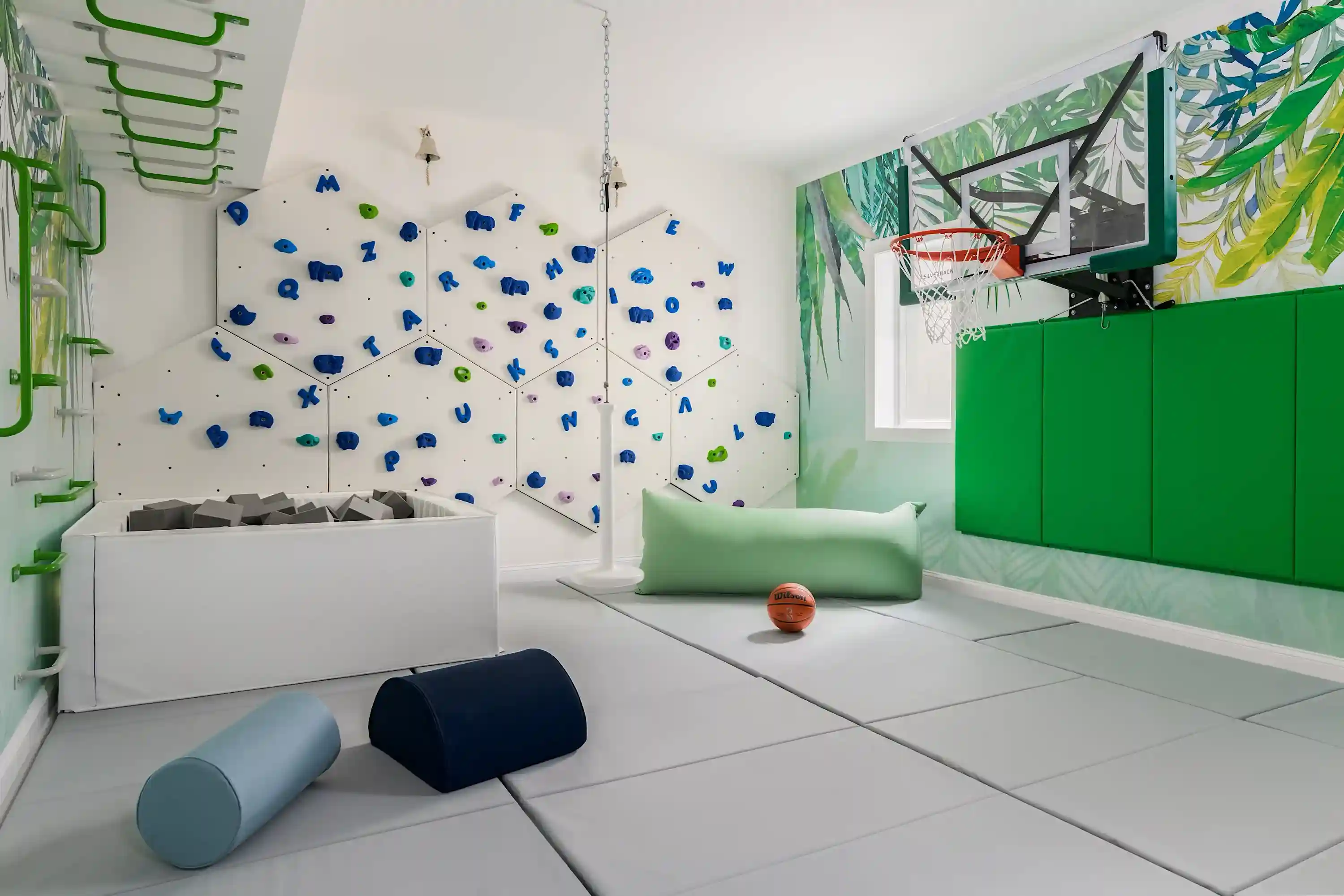 Indoor kids' playroom with climbing wall, foam pit, basketball hoop, padded floor mats, and colorful workout equipment.