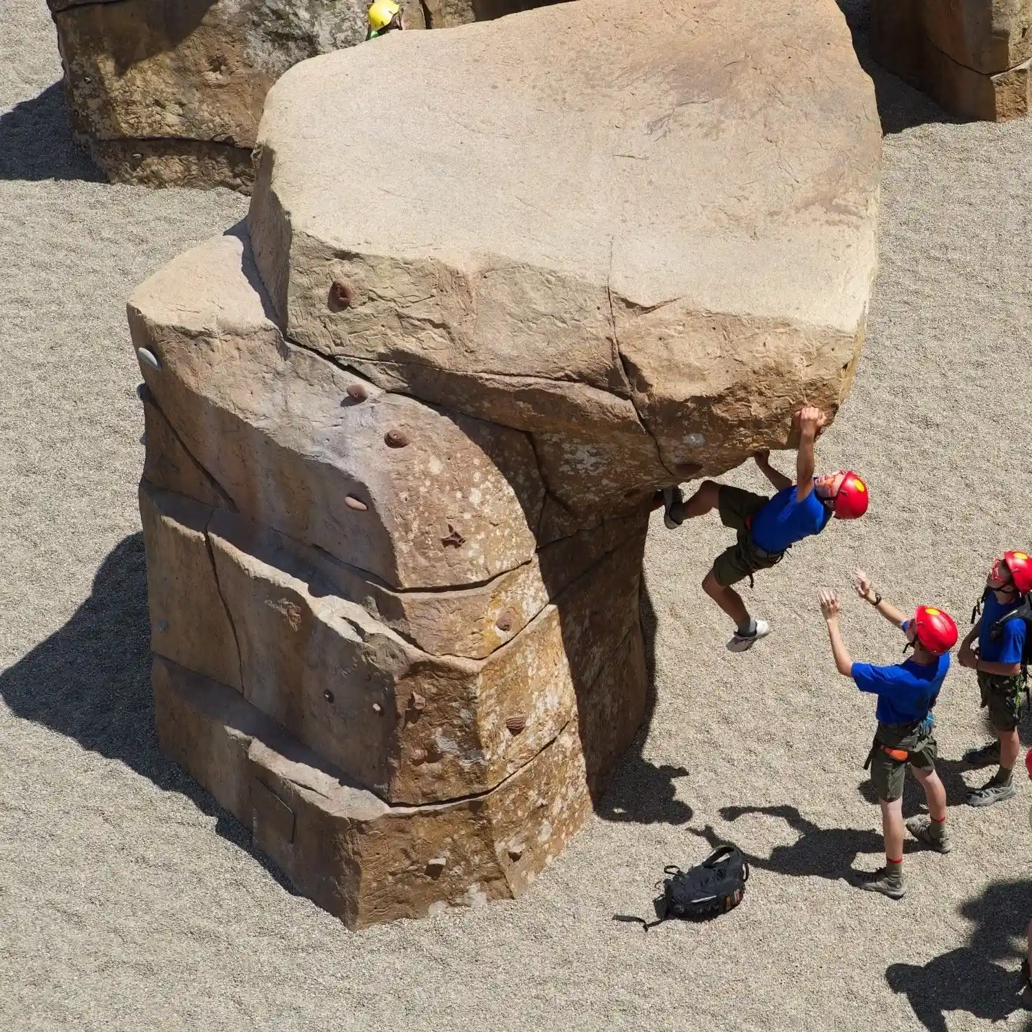 Person wearing a red helmet and climbing gear bouldering on a large artificial rock while two others in red helmets watch and spot him.