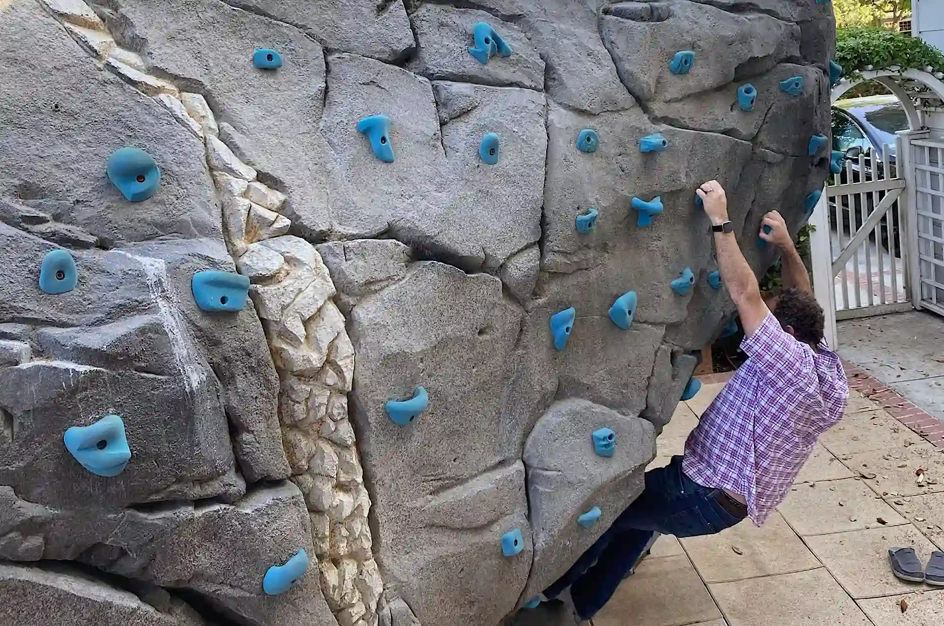 Man climbing on a gray backyard rock climbing wall with blue holds near a white gate and garden.