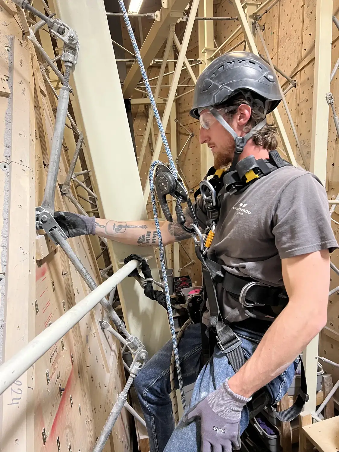 Worker wearing safety harness, helmet, gloves, and goggles inspecting a climbing wall indoors.