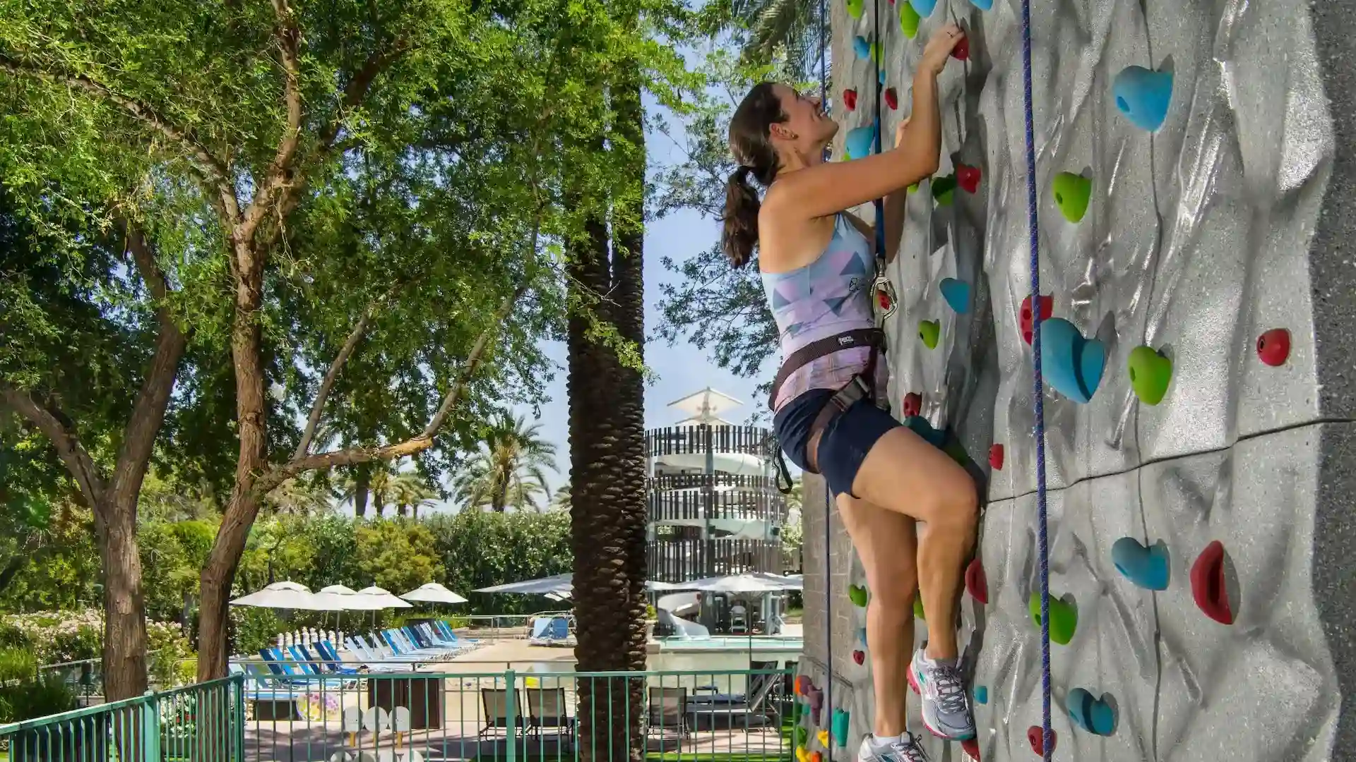 Woman climbing an outdoor rock climbing wall near a pool with lounge chairs and umbrellas in a resort setting.
