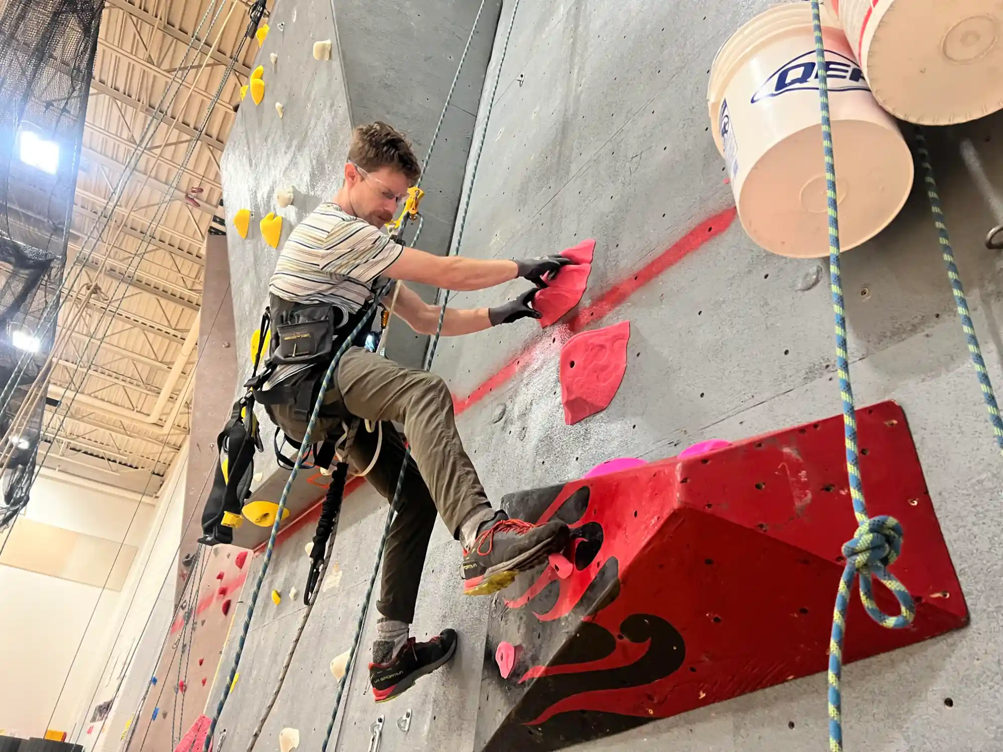 Man wearing climbing gear and gloves ascending an indoor rock climbing wall with colorful handholds.