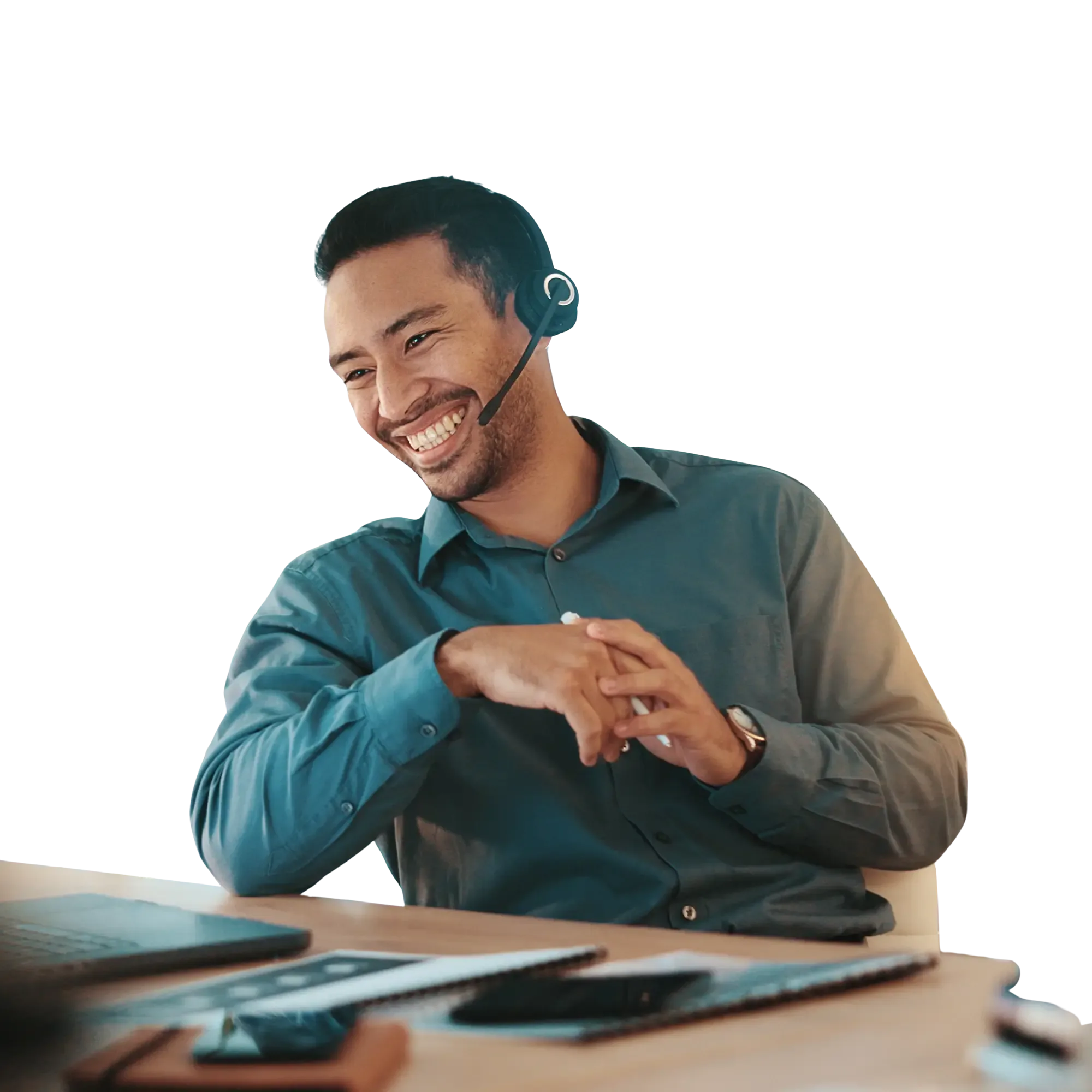Smiling man wearing a headset and blue shirt, sitting at a desk in an office with plants and shelves in the background.