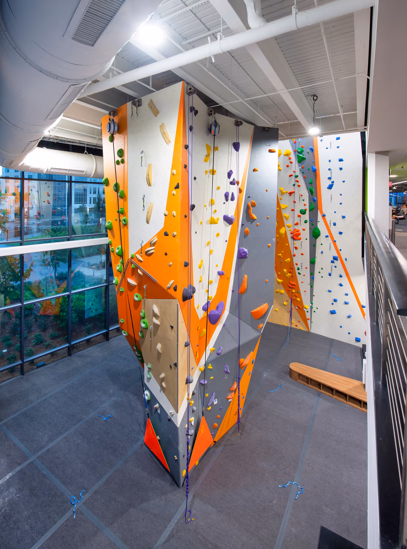 Indoor climbing gym with multi-colored climbing walls featuring orange, gray, and white panels and various climbing holds.