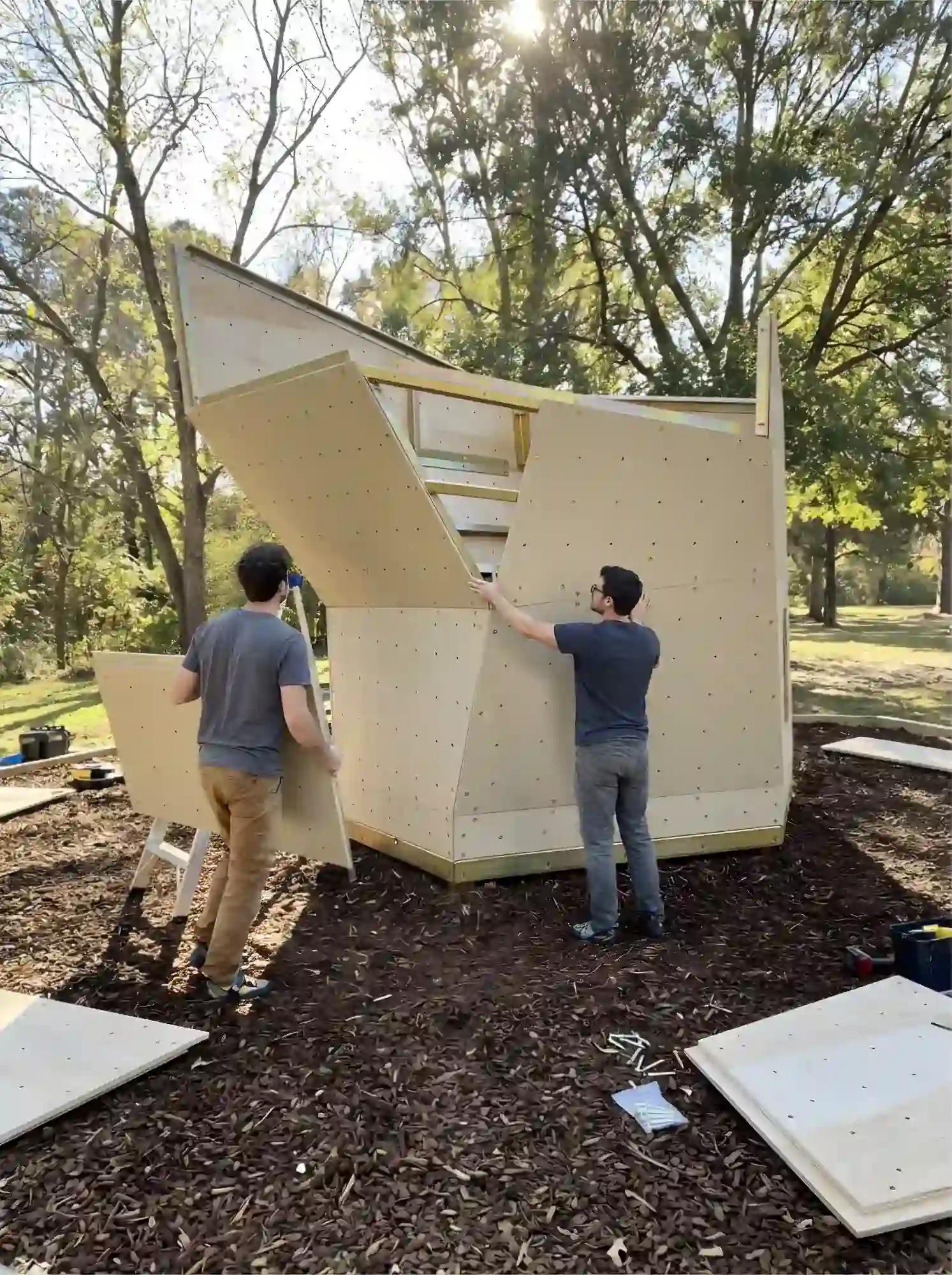Two men assembling a geometric wooden climbing wall structure outdoors in a wooded area with sunlight filtering through trees.