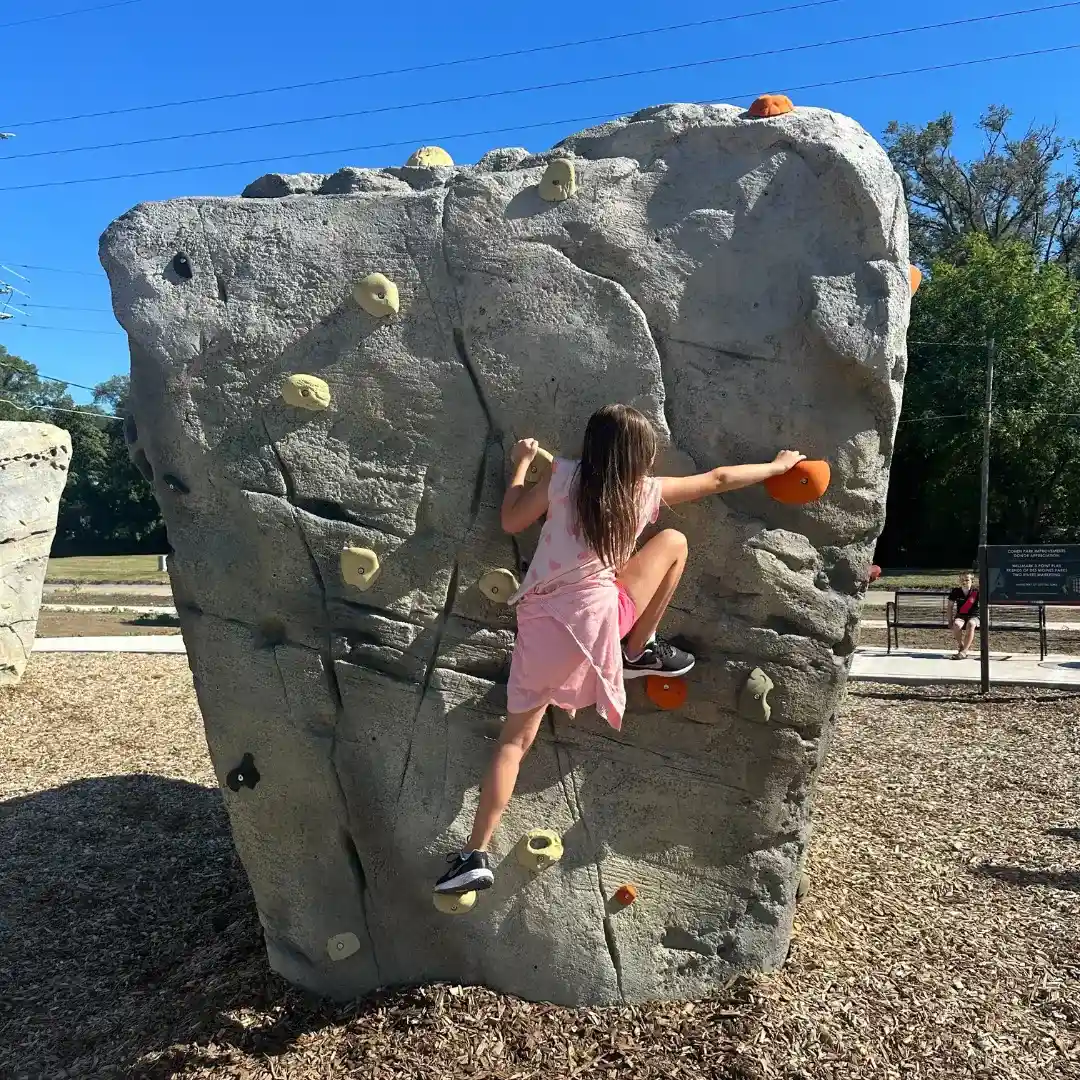 Young girl in a pink dress climbing a small outdoor rock climbing wall with orange and yellow holds.
