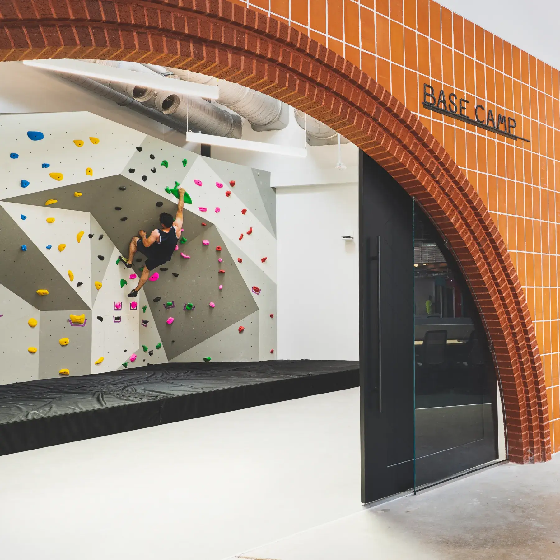 Indoor rock climbing gym with a climber on a bouldering wall inside a room with an arched brick entrance labeled Base Camp.