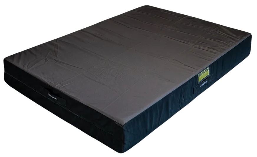 Black and gray crash pad on the floor in front of a climbing wall with colorful holds.