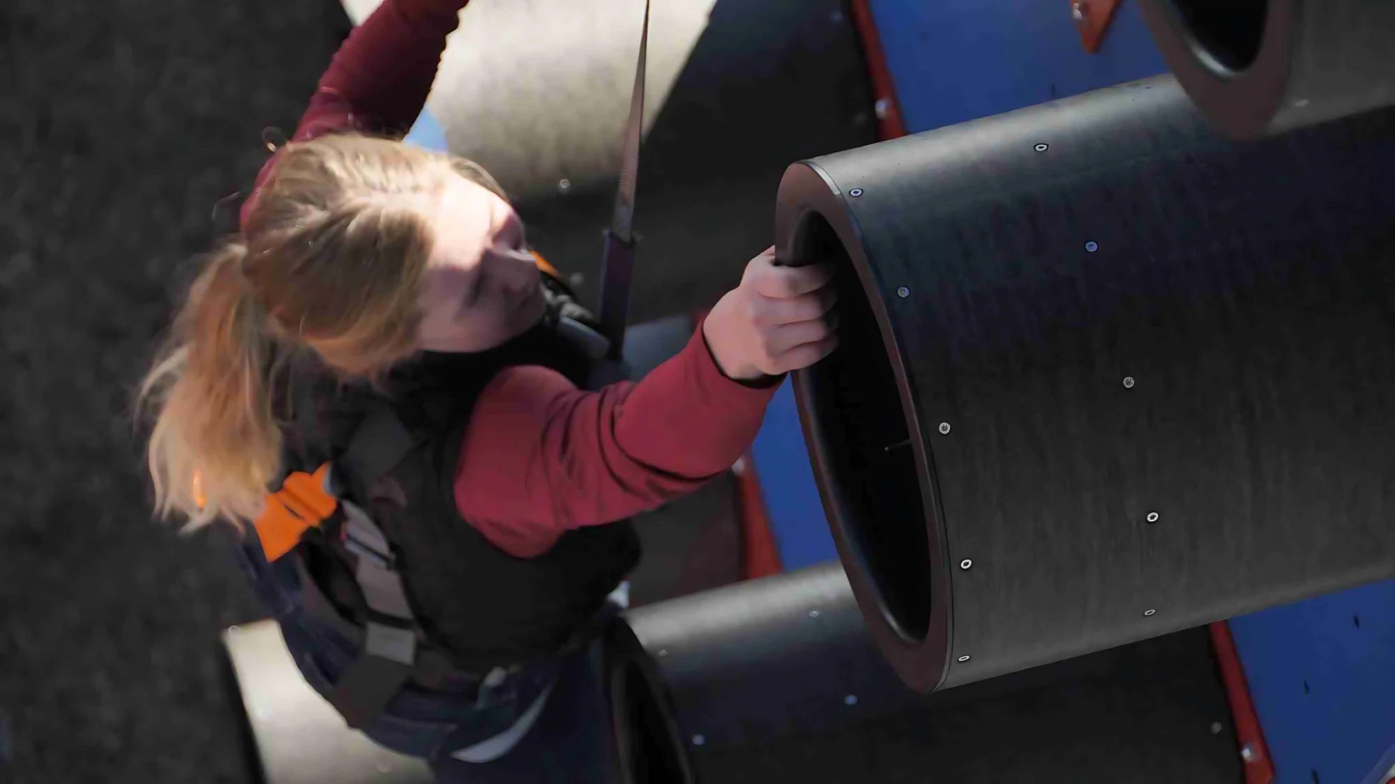 Person climbing a wall using large cylindrical grips with a safety harness attached.