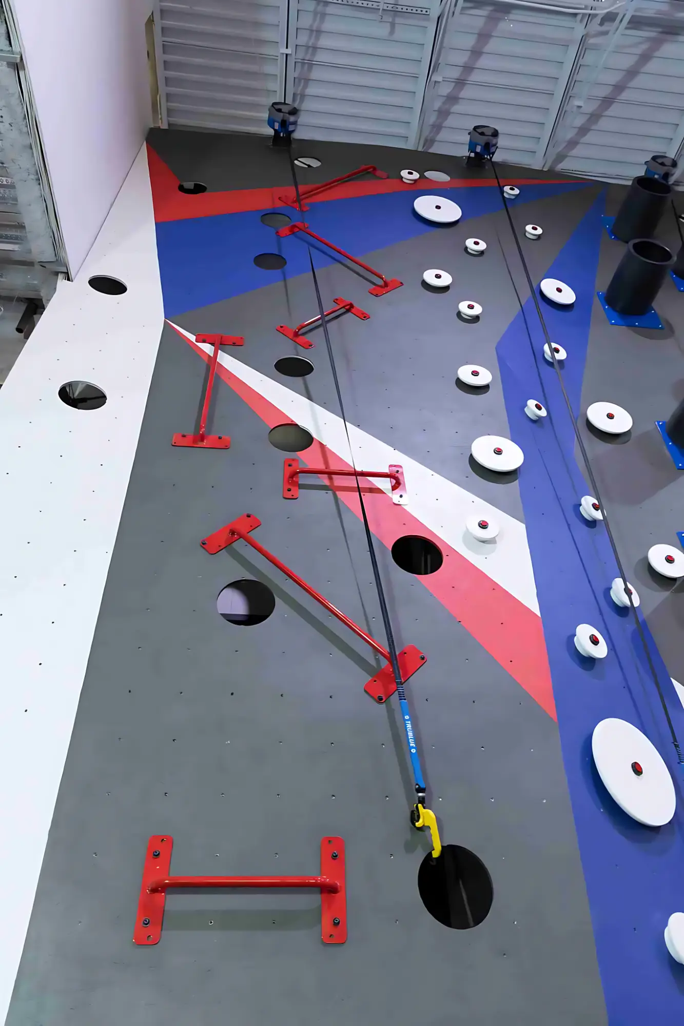 Four children climbing a black outdoor bouldering wall with colorful holds and safety mats underneath at a playground.