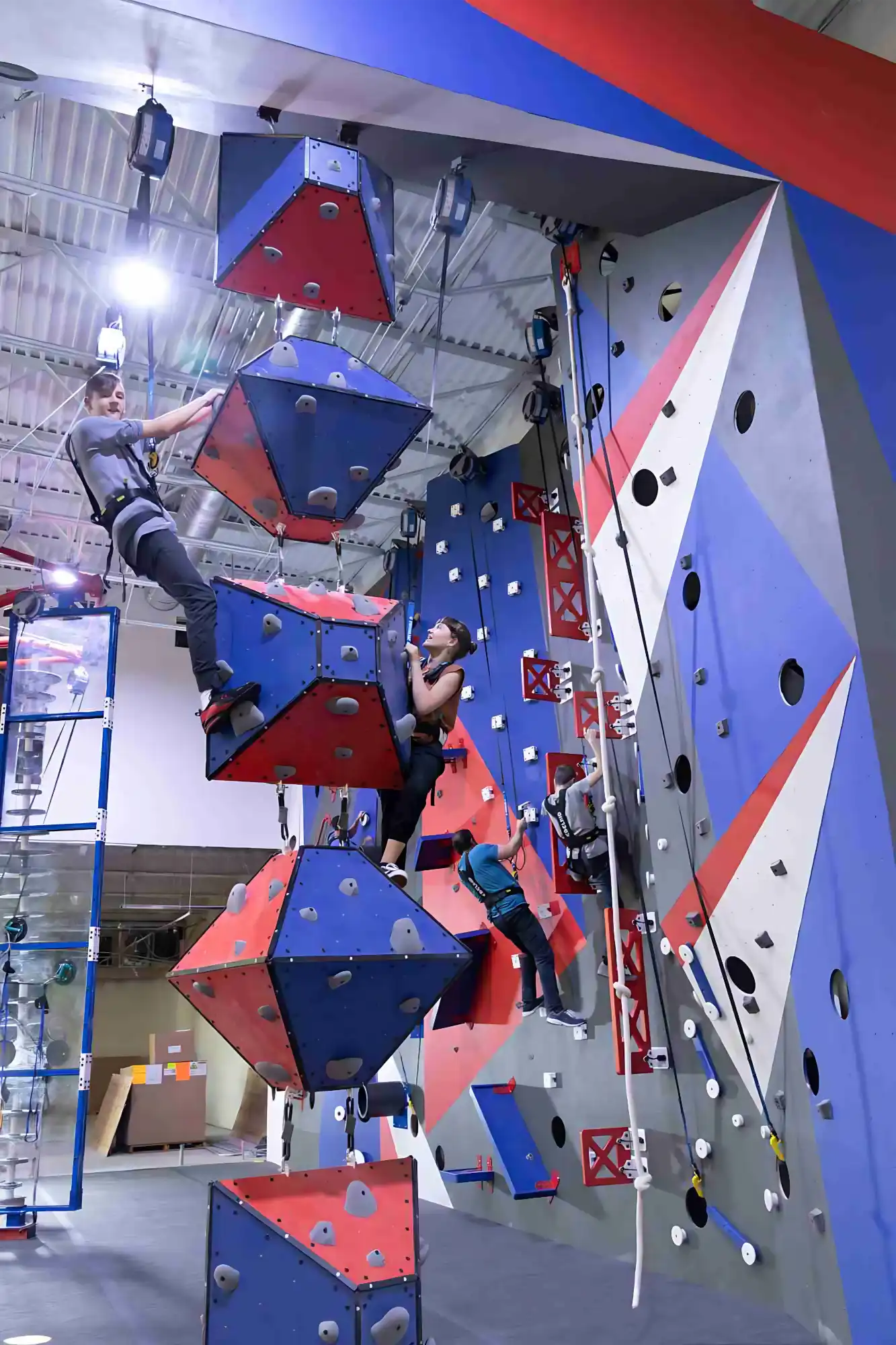 People climbing on three outdoor bouldering walls with red, yellow, and blue holds in a park setting.