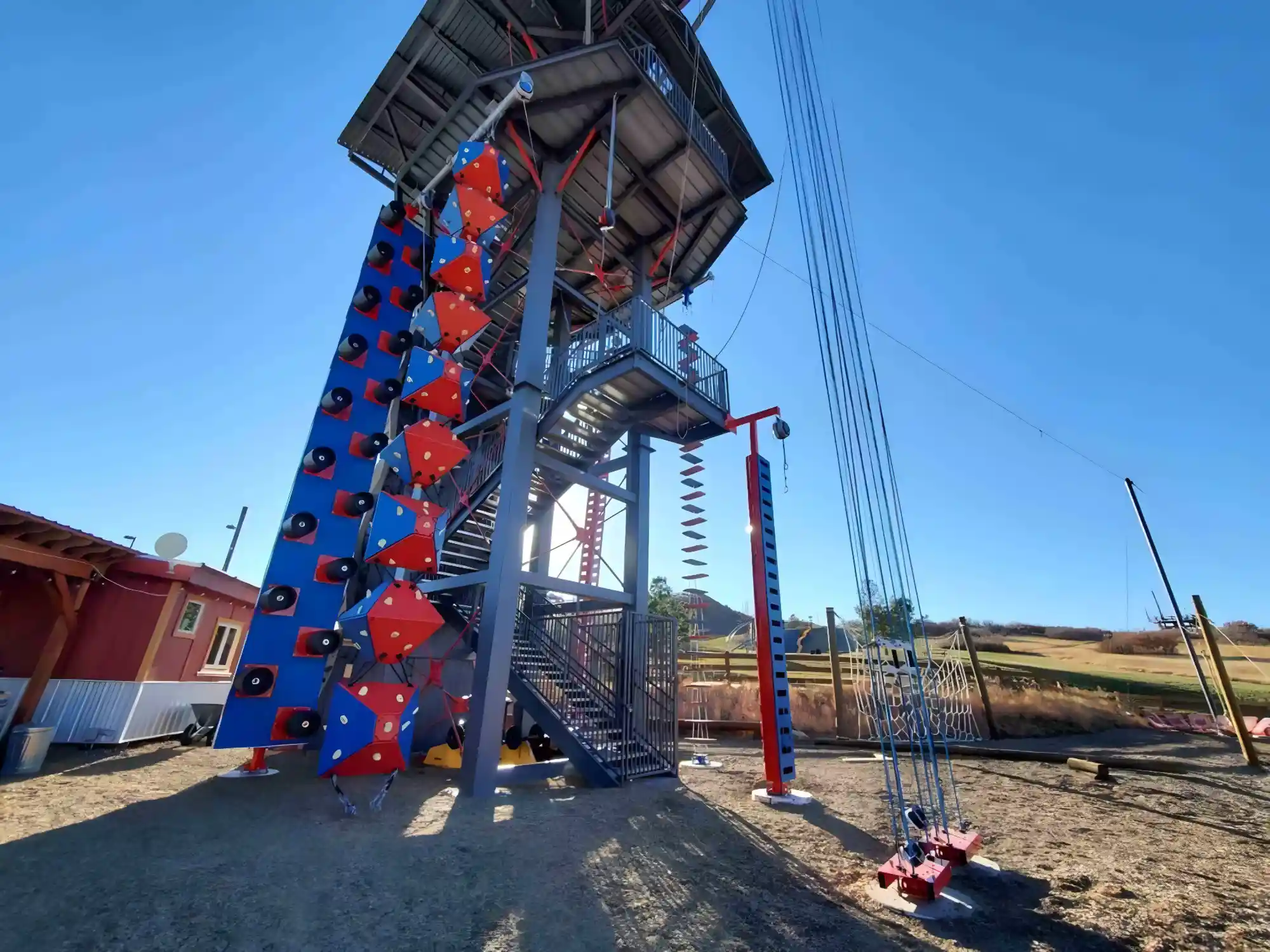 Outdoor climbing tower with blue and red climbing holds, ropes, and platforms under clear blue sky.
