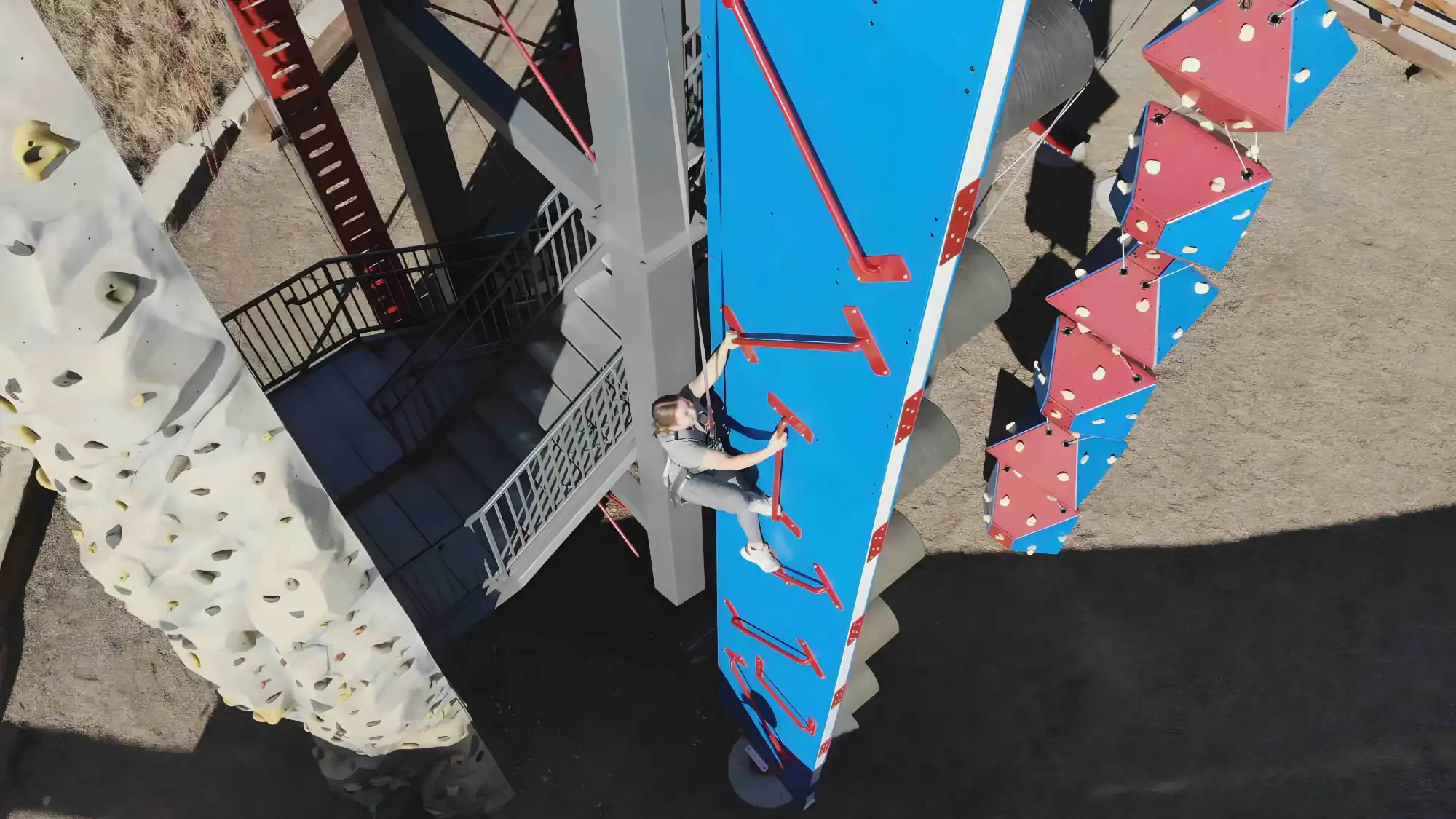 Person climbing a blue and red vertical wall climber structure outdoors next to a white climbing wall.