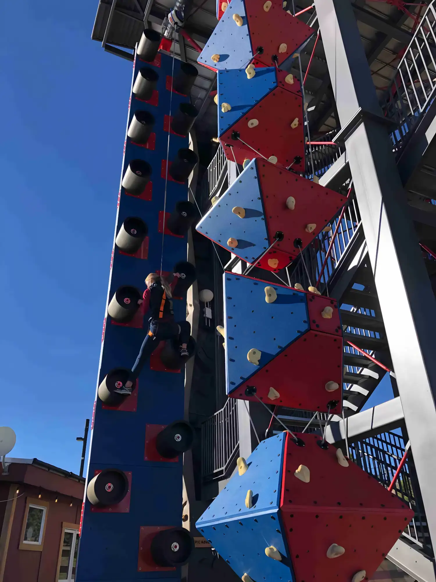 Person climbing a blue wall with large cylindrical holds at an outdoor climbing gym.