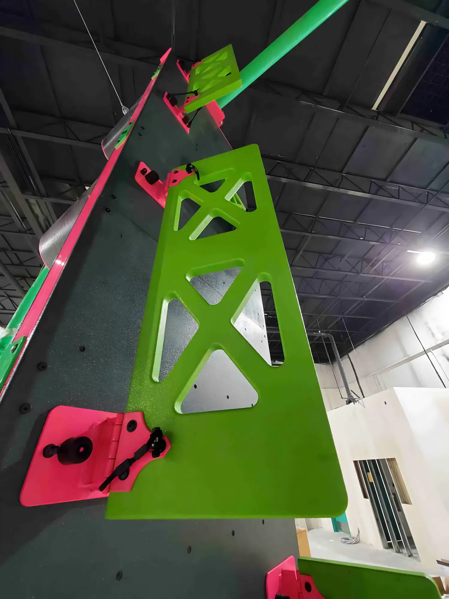 Bright green and pink climbing holds attached to a tall indoor climbing wall under a black ceiling.