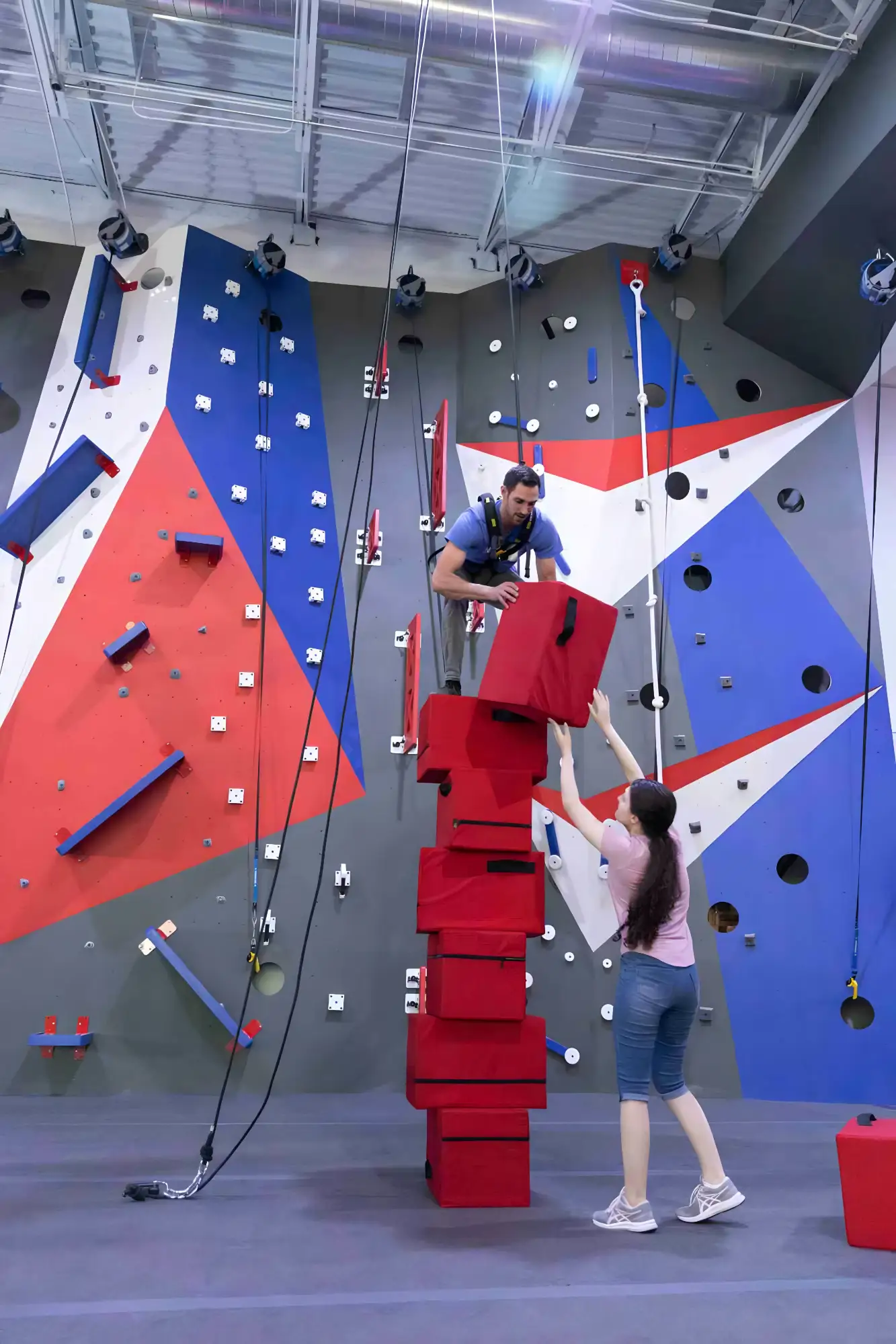 Man climbing and stacking red foam blocks while woman assists in an indoor climbing gym.