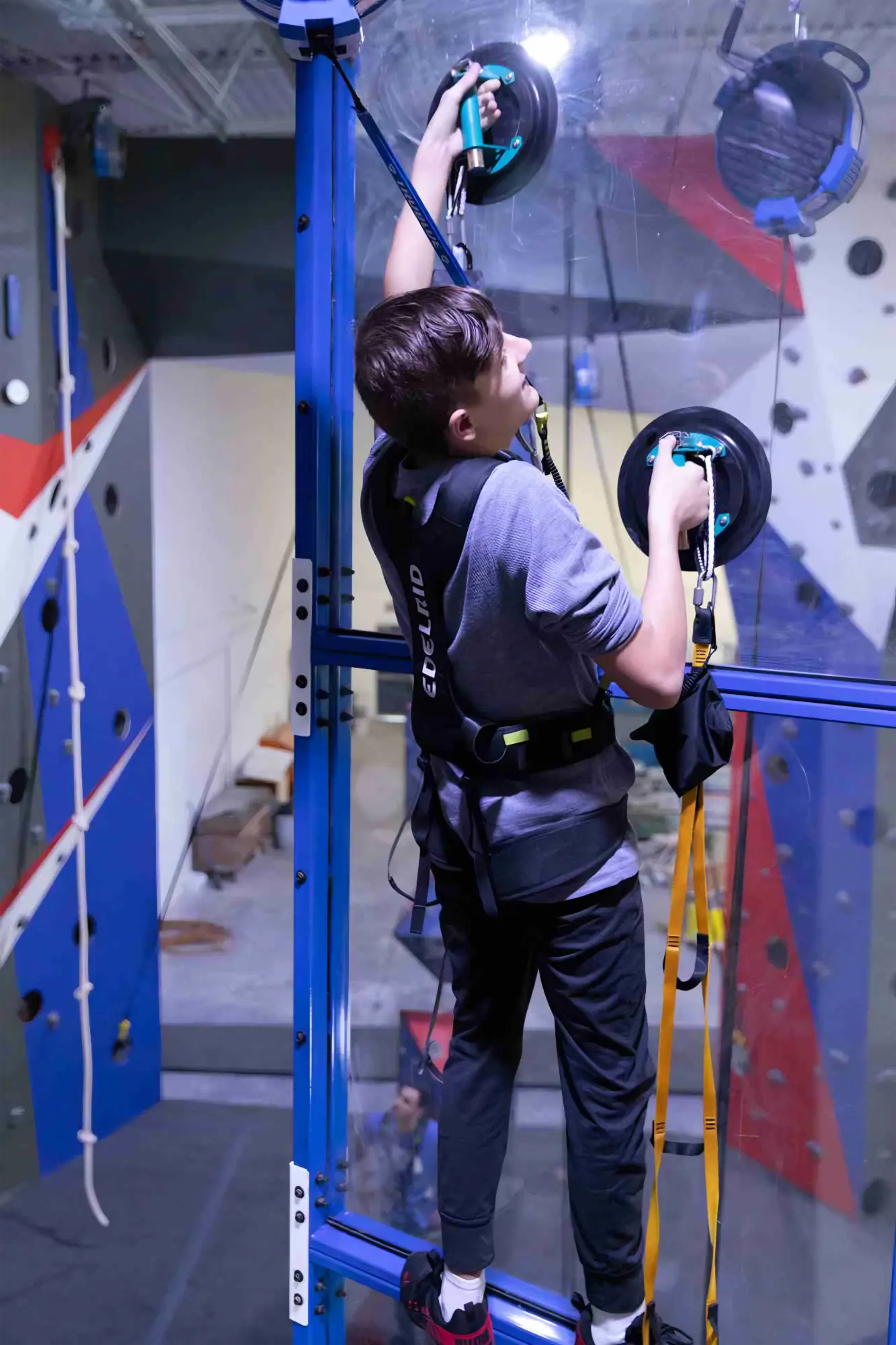 Young person indoor climbing on vertical glass wall with suction cup grips and safety harness.