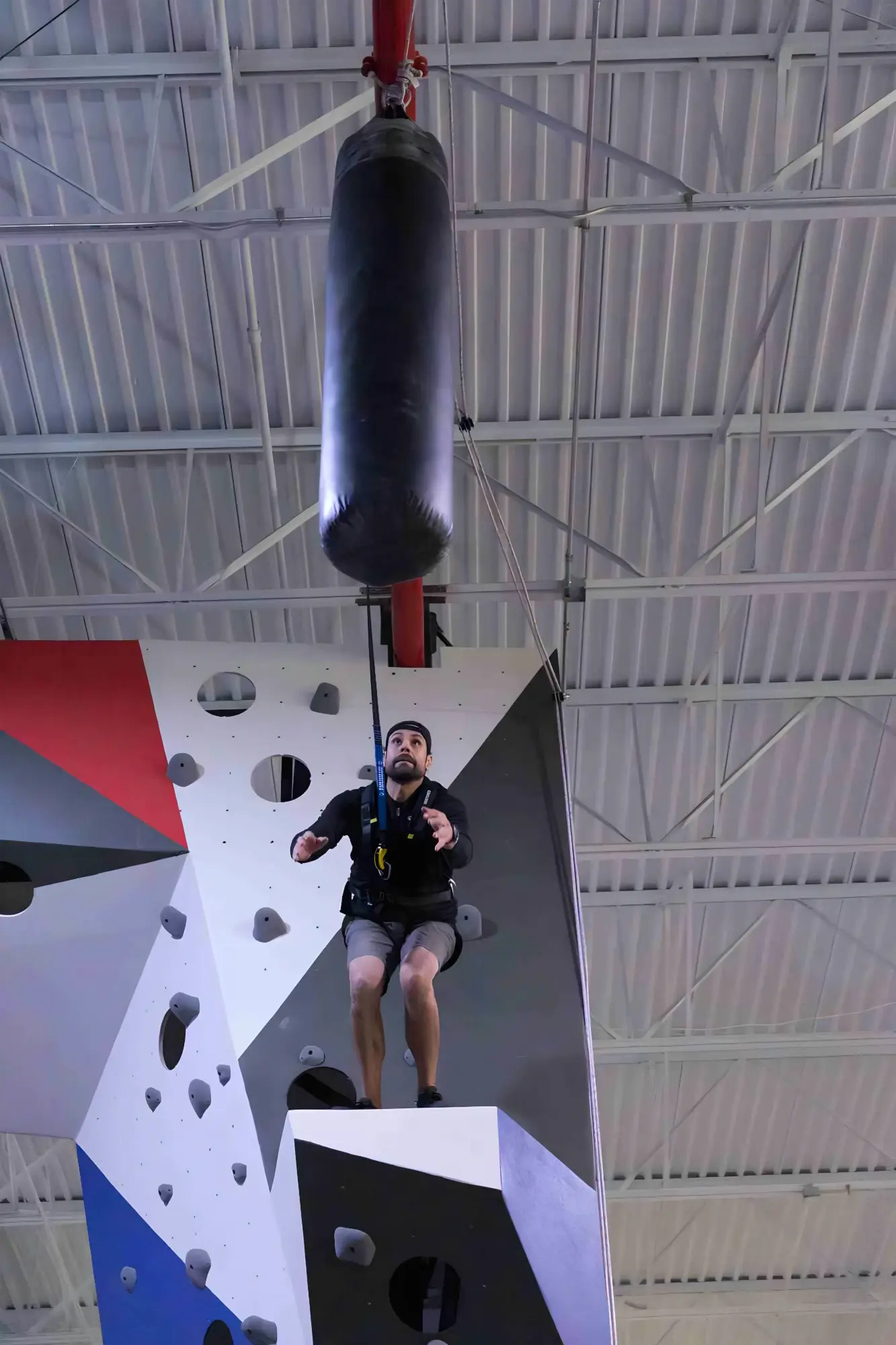 Man in harness jumping from climbing wall platform toward a hanging punching bag in gym.