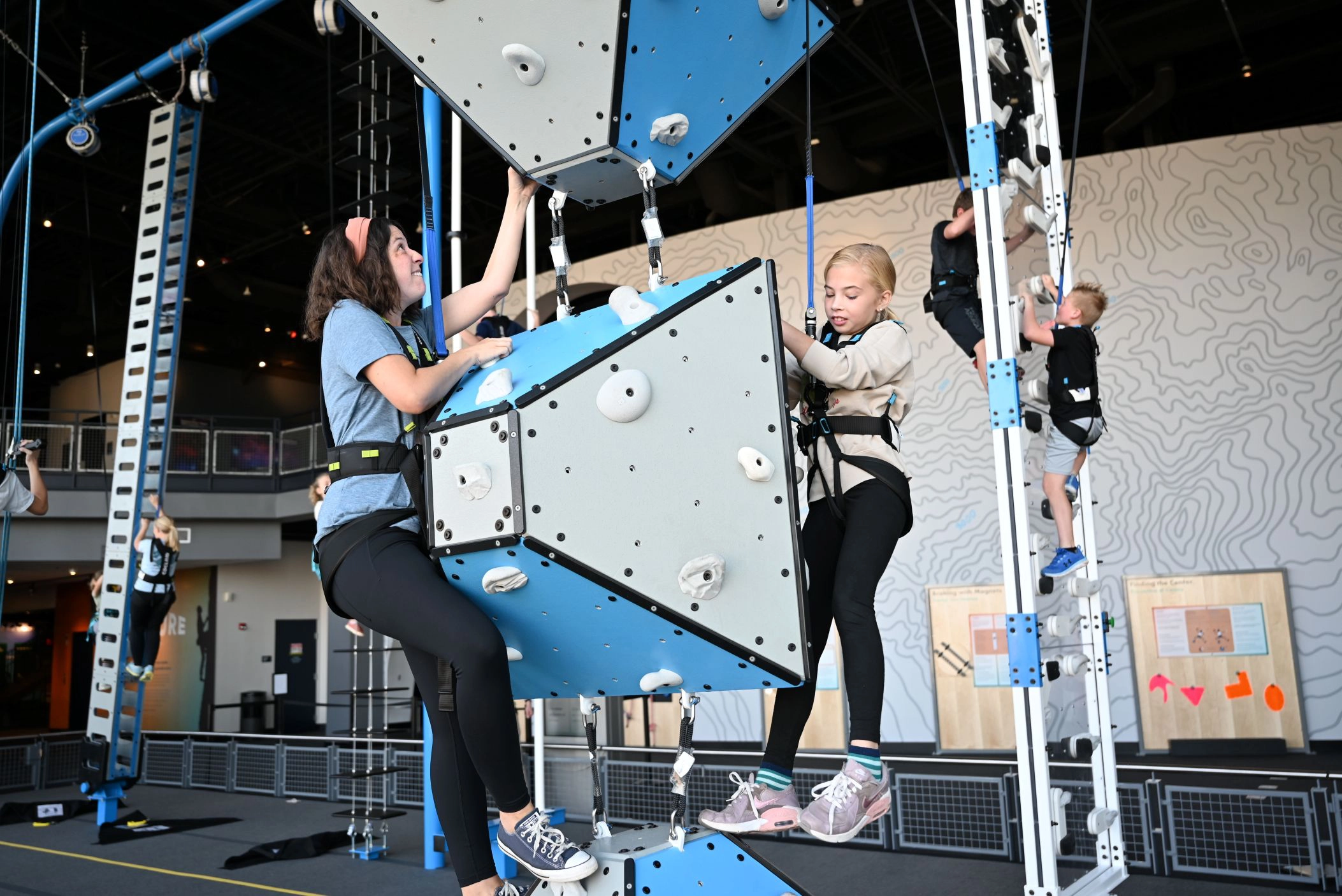 Two girls climbing a blue and gray indoor rock climbing structure wearing harnesses.