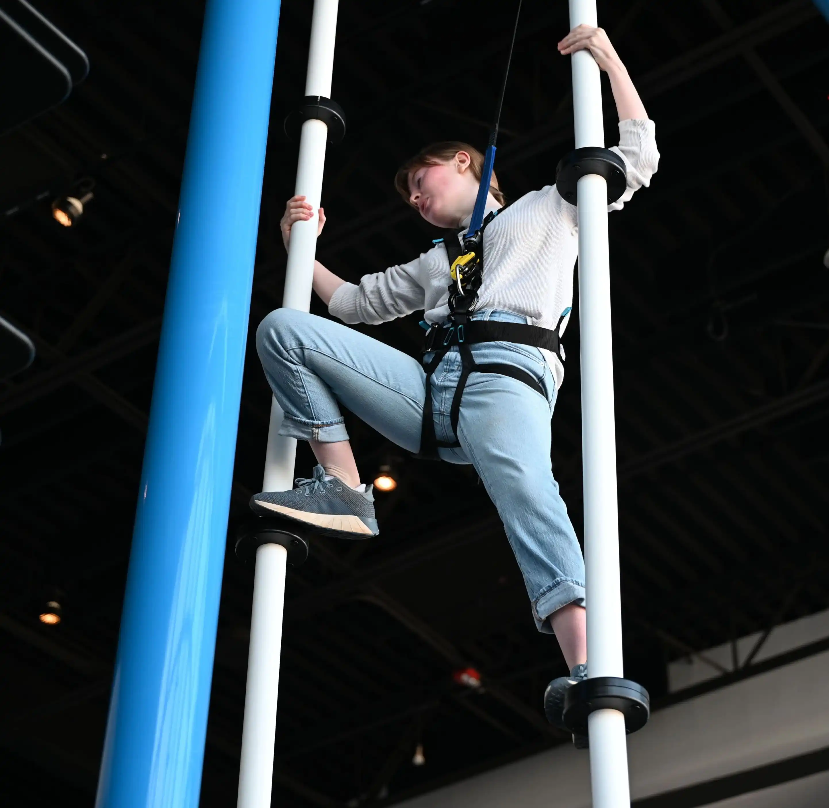 Person wearing a harness climbing two vertical poles indoors with black ceiling beams above.