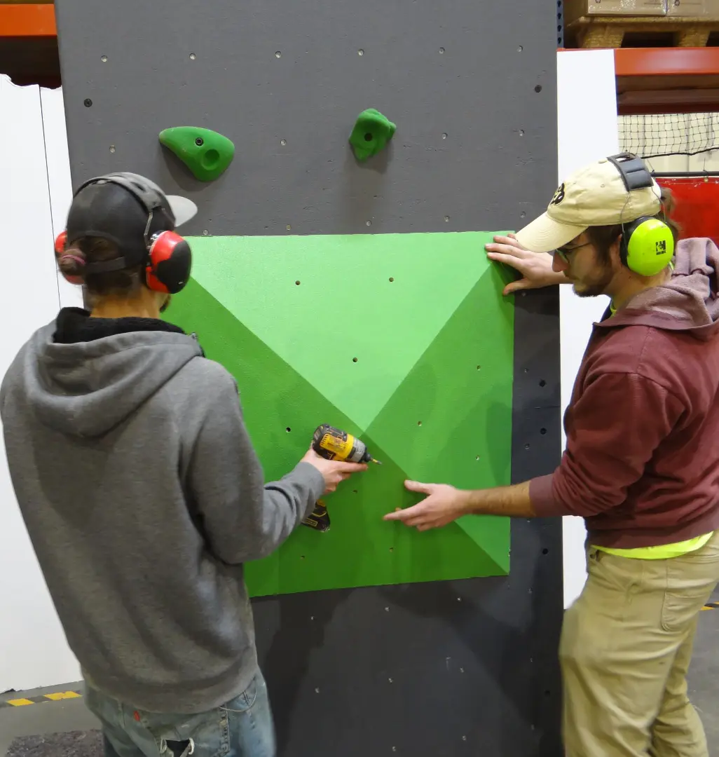 Two people wearing ear protection install a green panel on a gray climbing wall indoors.