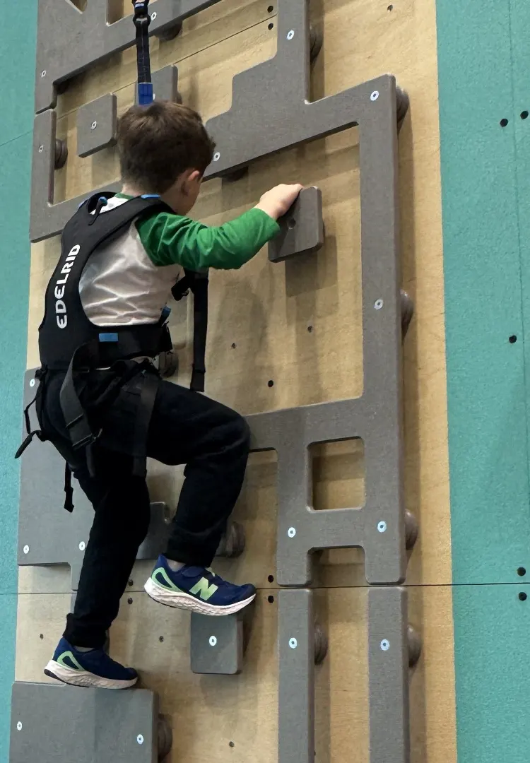 Young child wearing climbing harness ascending an indoor climbing wall with large gray holds.
