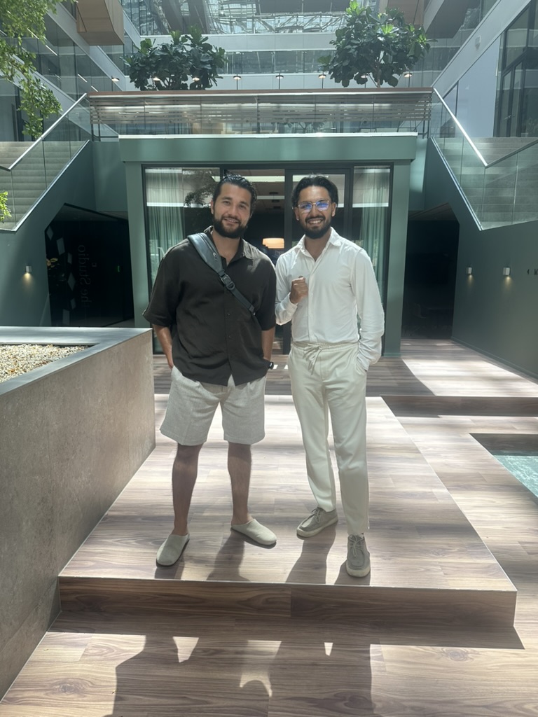 Two men standing indoors on a wooden platform with modern glass and plant decor in the background, one wearing a dark shirt and shorts, the other in a white shirt and light pants.