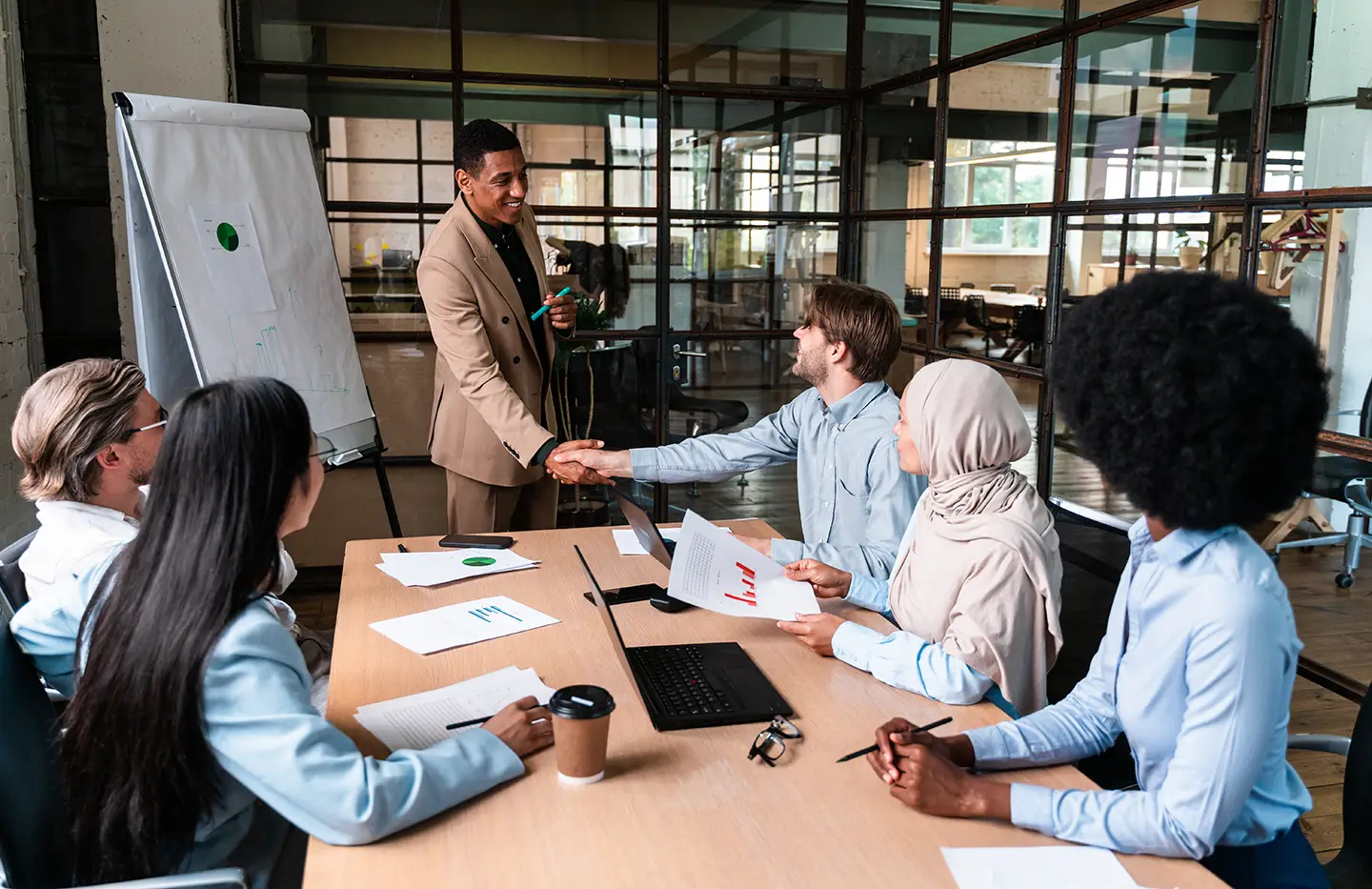 A business meeting with seven professionals engaging at a conference table, one person shaking hands with another, while others look on with documents and a laptop in front of them. A whiteboard is in the background.