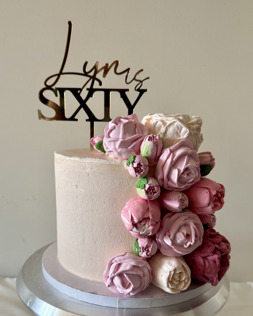 Cream-colored cake decorated with pink and white buttercream flowers and a gold 'Lynus Sixty' topper.