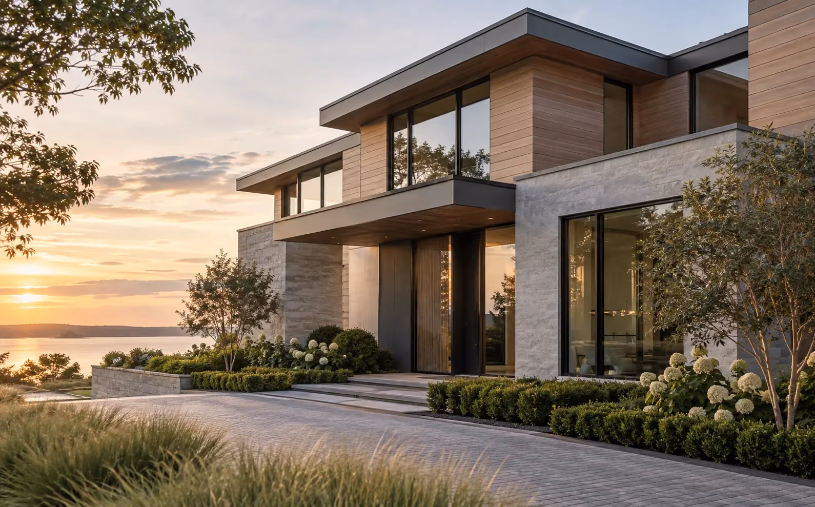 Modern luxury home with clean rooflines, subtle metal detailing, stone and wood siding, and large windows overlooking the water at sunset.