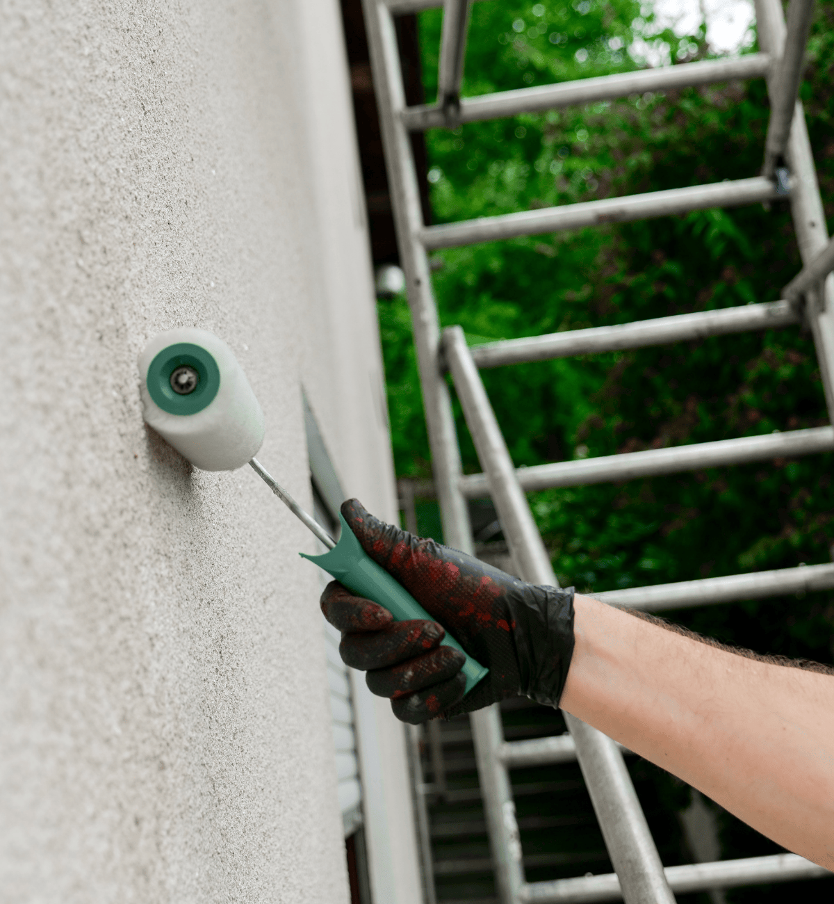 Person wearing a black glove painting a textured exterior wall with a small paint roller near a ladder.