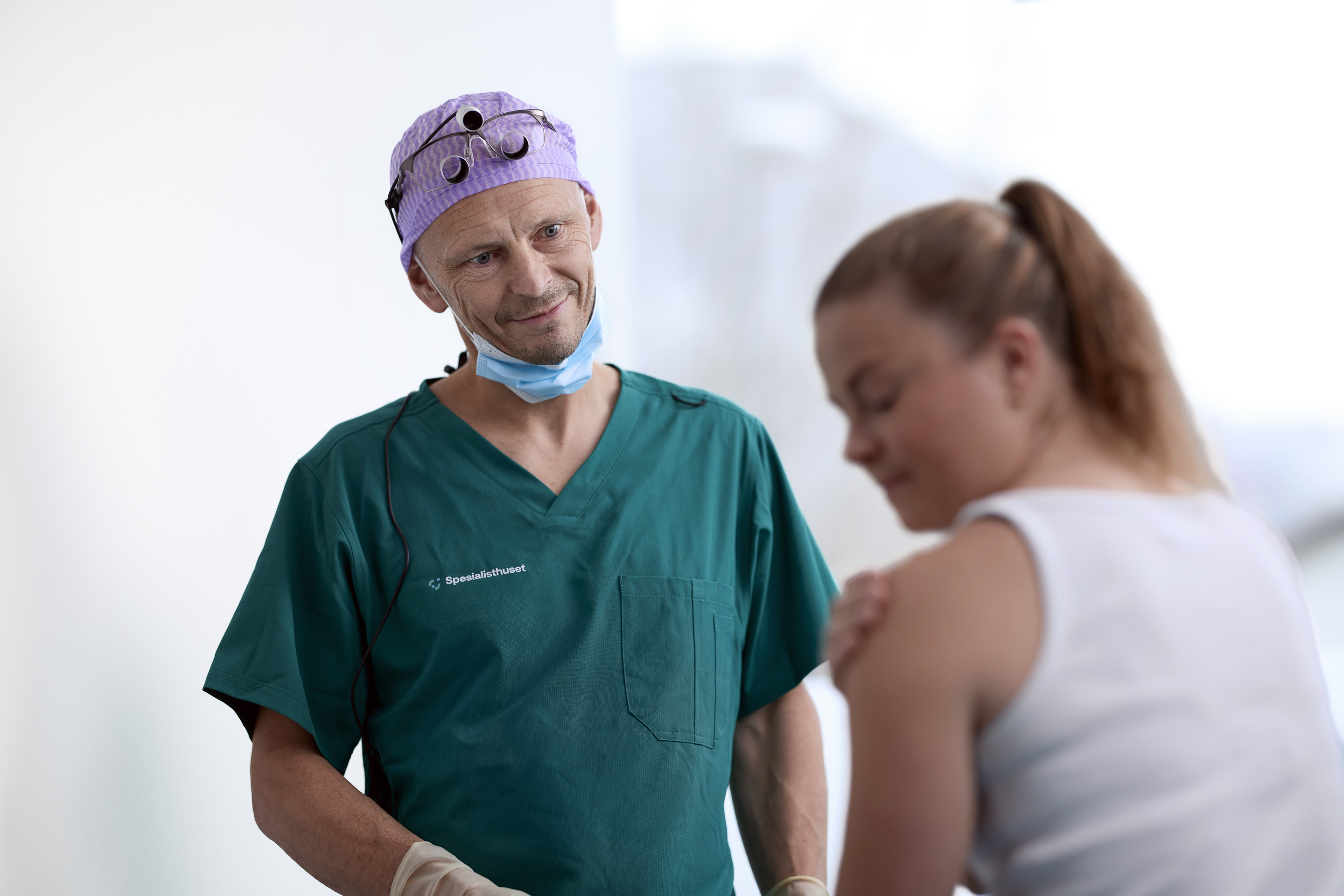 Lege i grønne scrubs og kirurgisk hette ser omsorgsfullt på en kvinne som klør på armen sin.