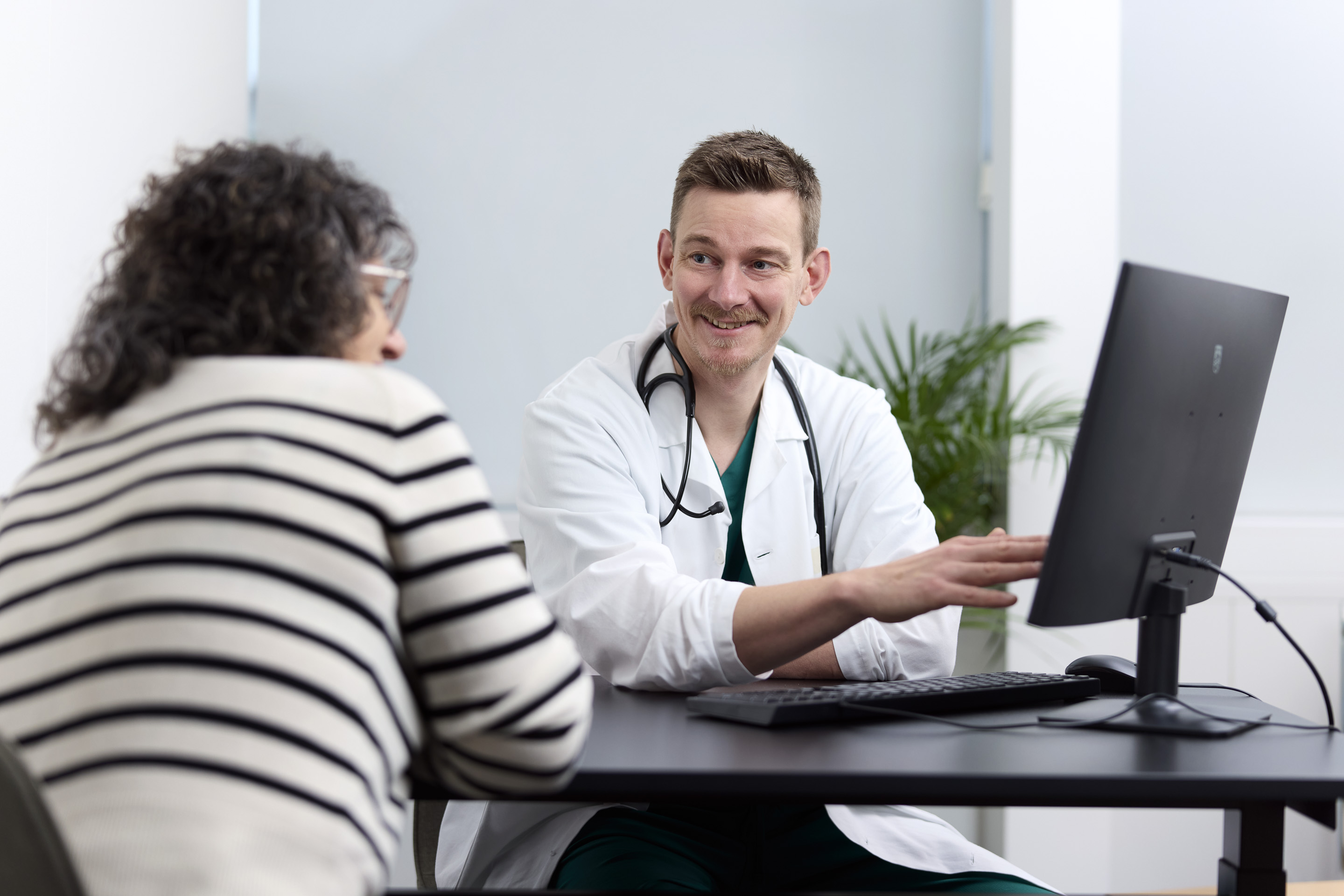 Lege i hvit frakk med stetoskop diskuterer noe på dataskjermen med en kvinnelig pasient i stripete genser.