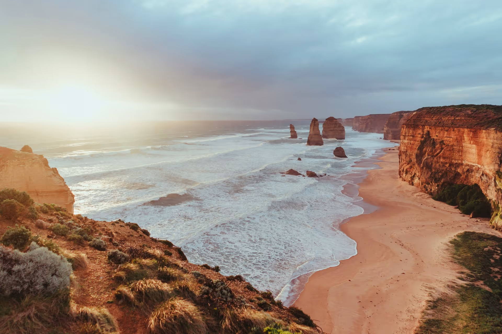 Sunset over ocean waves crashing against tall sandstone cliffs and rock formations along a sandy beach.