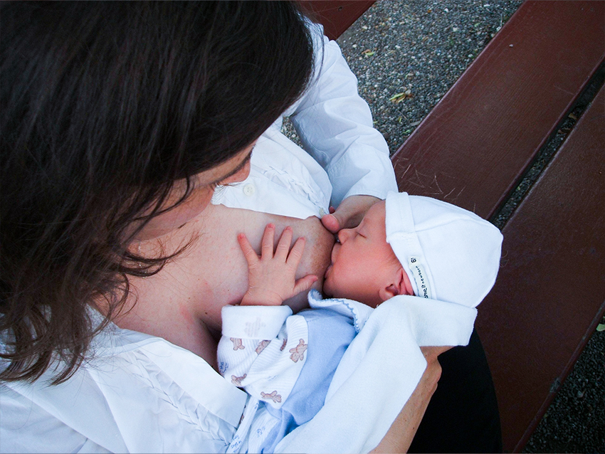 Mutter stillt ihr Baby, das eine weisse Mütze und einen blauen Strampler trägt, auf einer Parkbank.