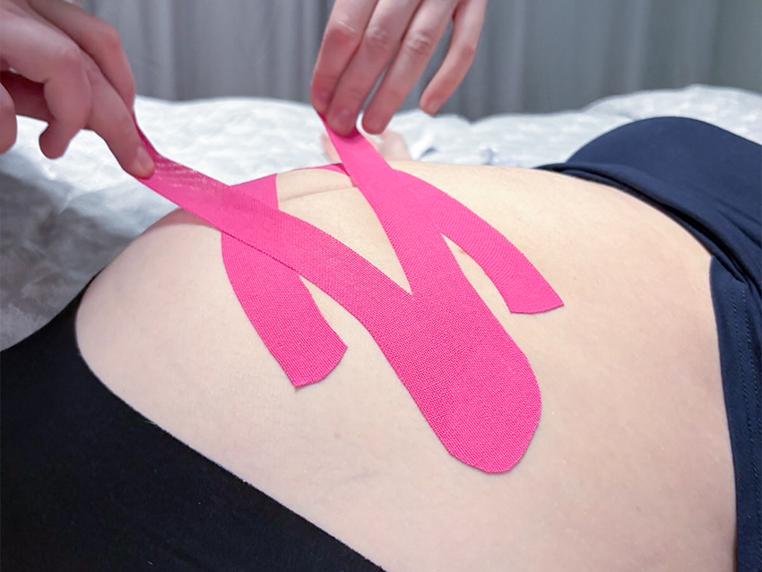 Hände, die rosa Kinesio-Tape in Form eines Y auf den Rücken einer liegenden Person kleben.