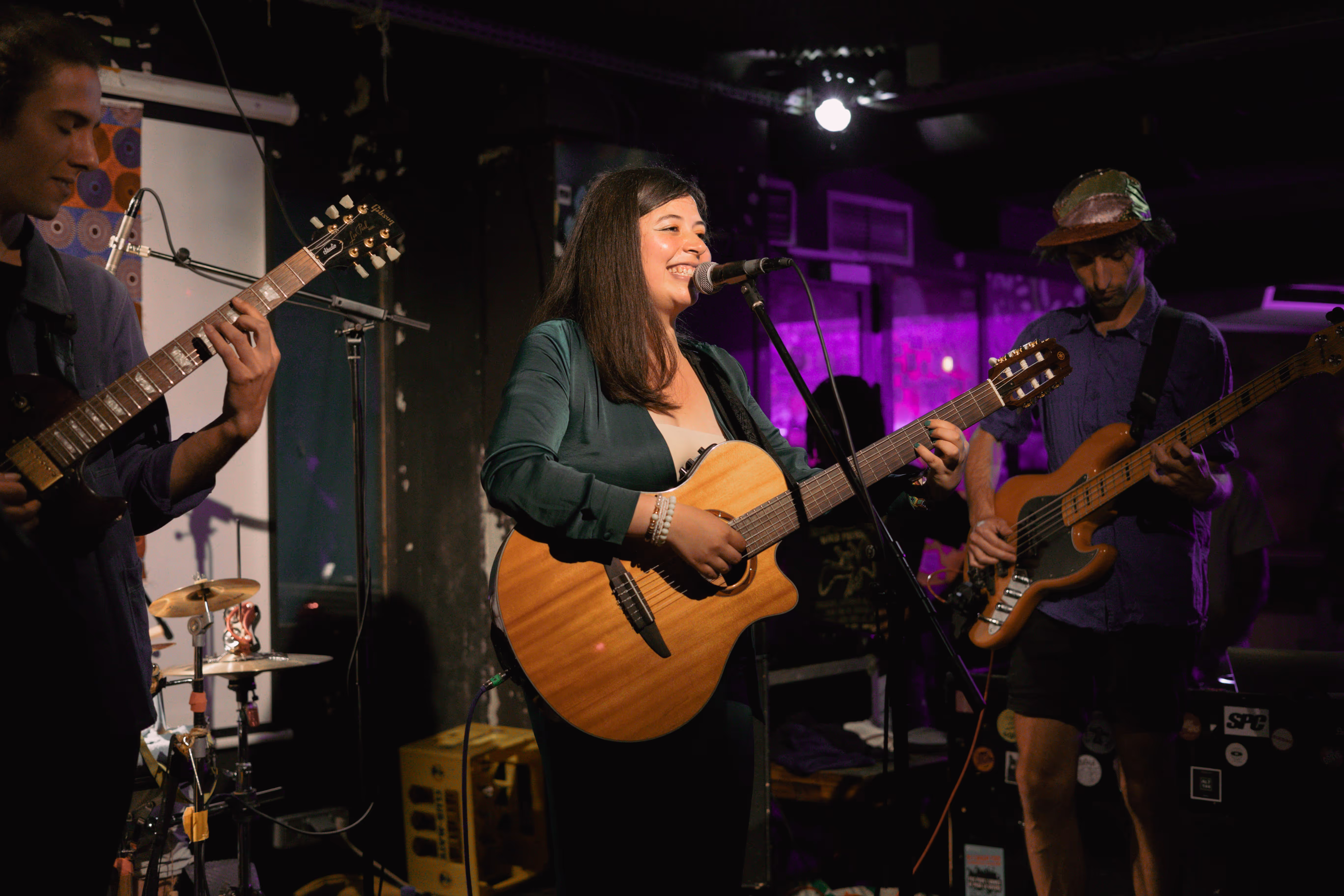 Chanteuse souriante jouant de la guitare acoustique dans un groupe avec deux musiciens sur scène.