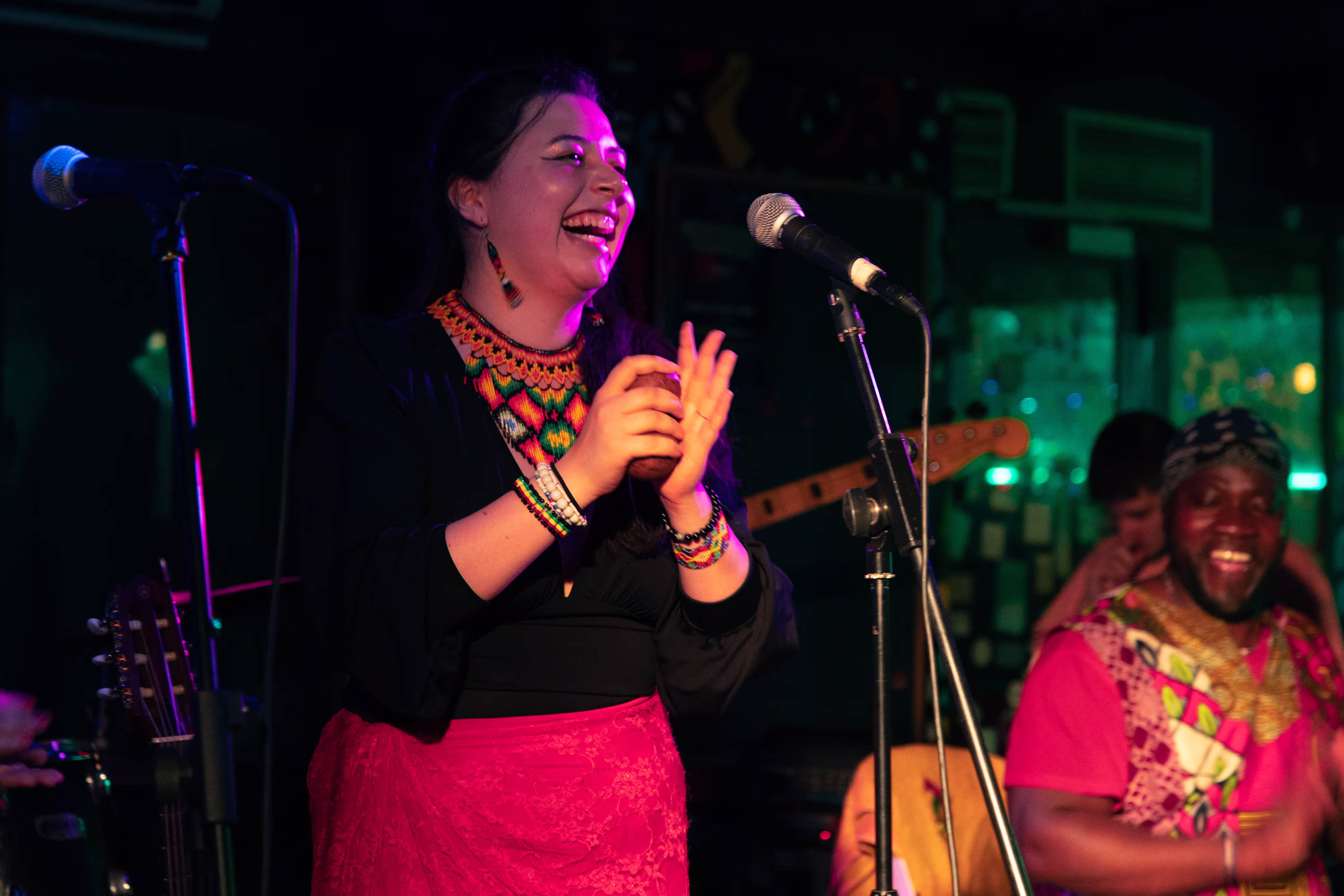 Une femme souriante en tenue colorée applaudit près d'un micro lors d'une performance musicale.