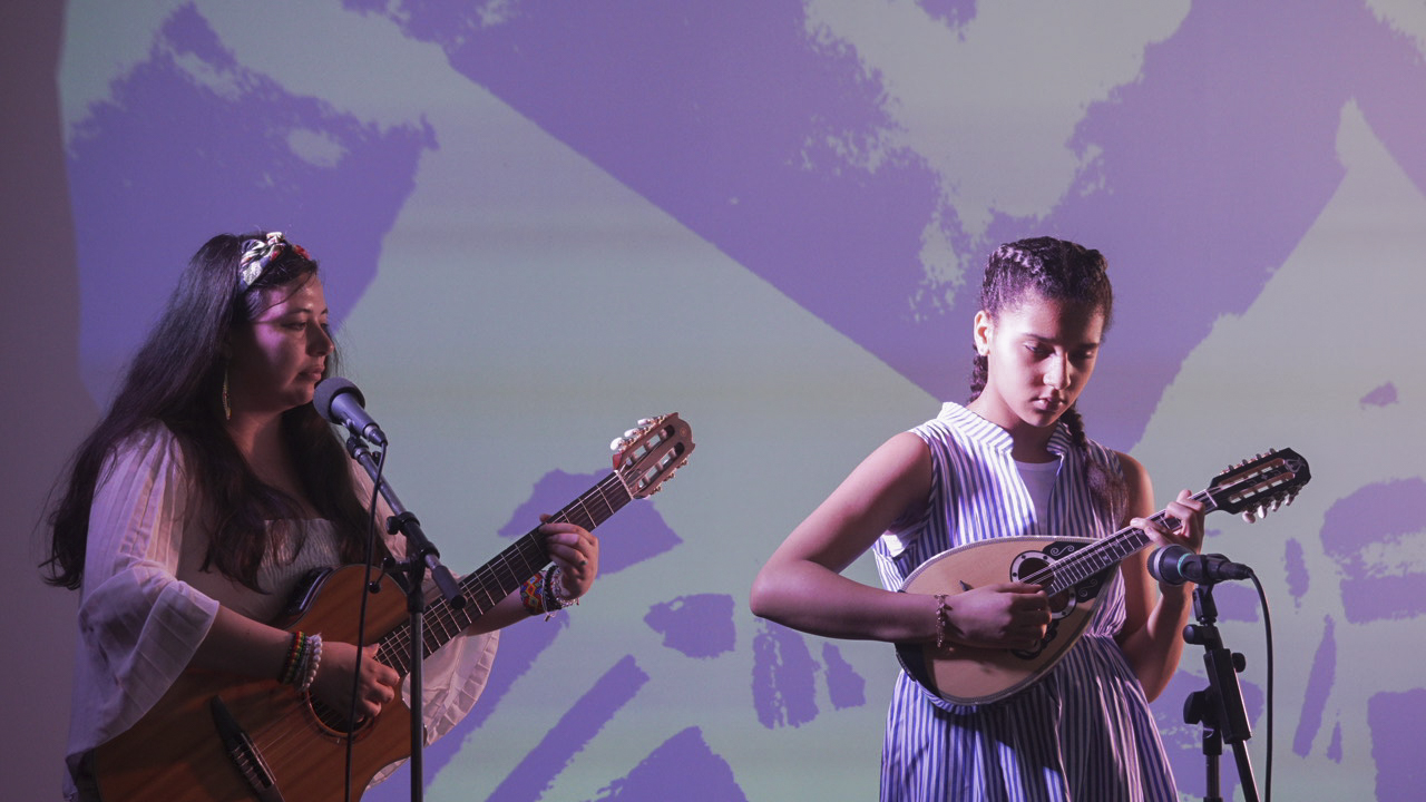 Deux femmes jouent de la guitare et du mandoline sur scène avec un micro et fond violet abstrait.
