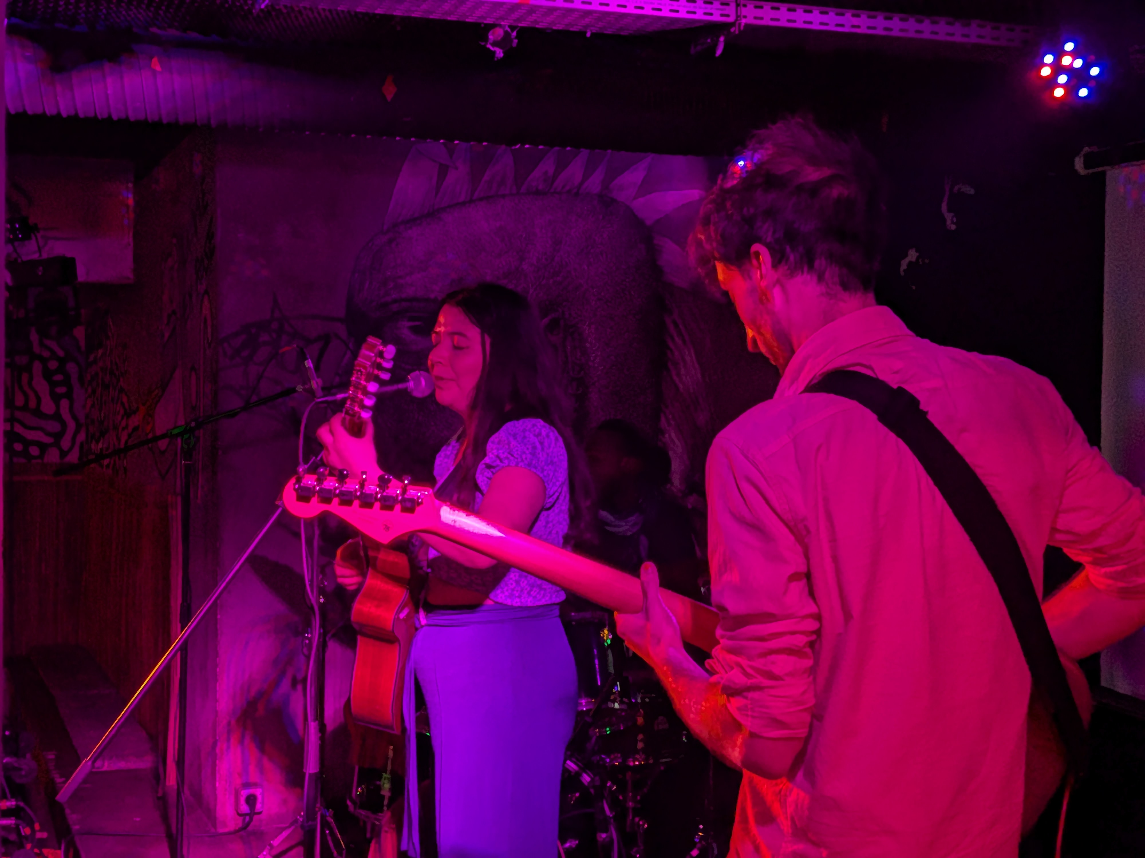Deux musiciens jouent de la guitare et chantent sur scène sous un éclairage violet.