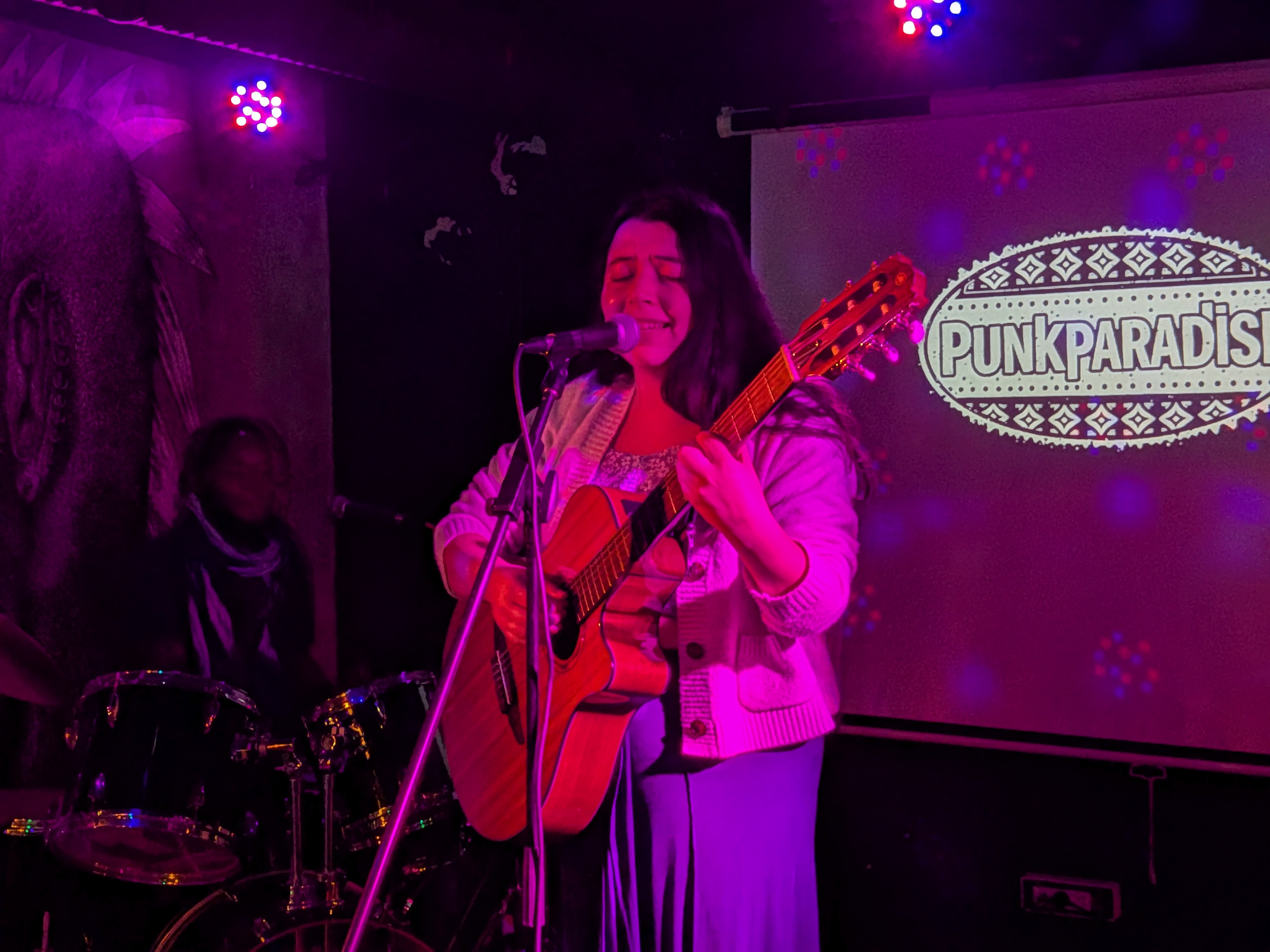 Femme chantant et jouant de la guitare sur scène avec un batteur et un logo PunkParadise en arrière-plan.