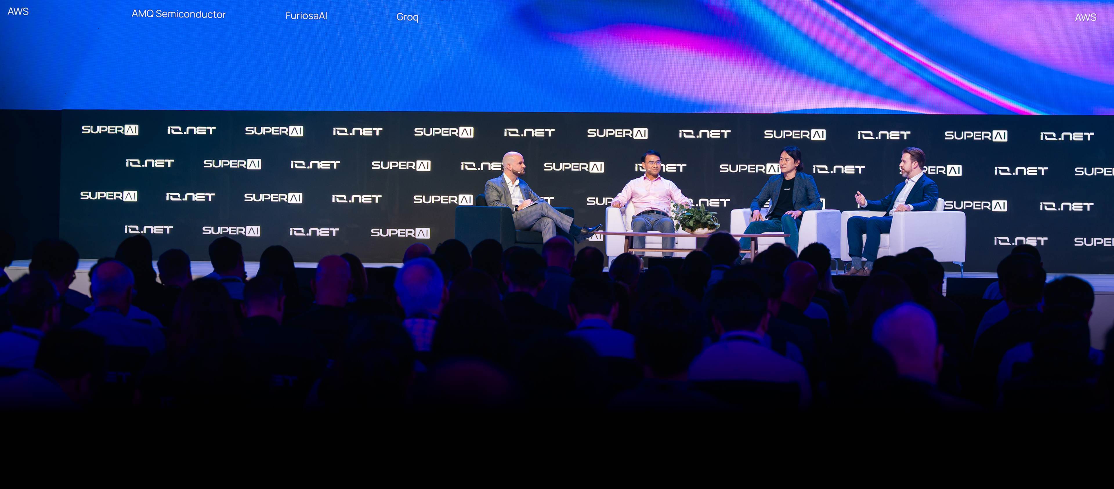 Three men seated on a stage panel discussion with SUPERAI and IQ.NET logos on a black backdrop and an audience in the foreground.