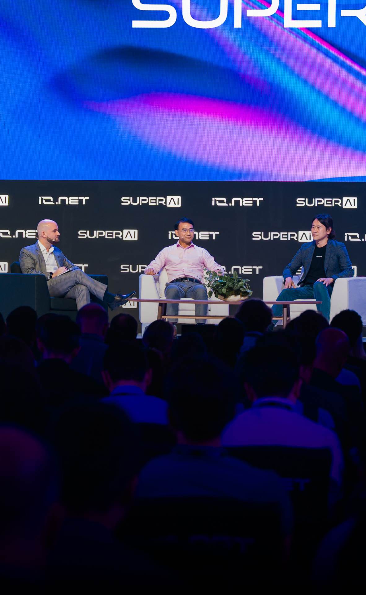 Three men seated on a stage panel discussion with SUPERAI and IQ.NET logos on a black backdrop and an audience in the foreground.
