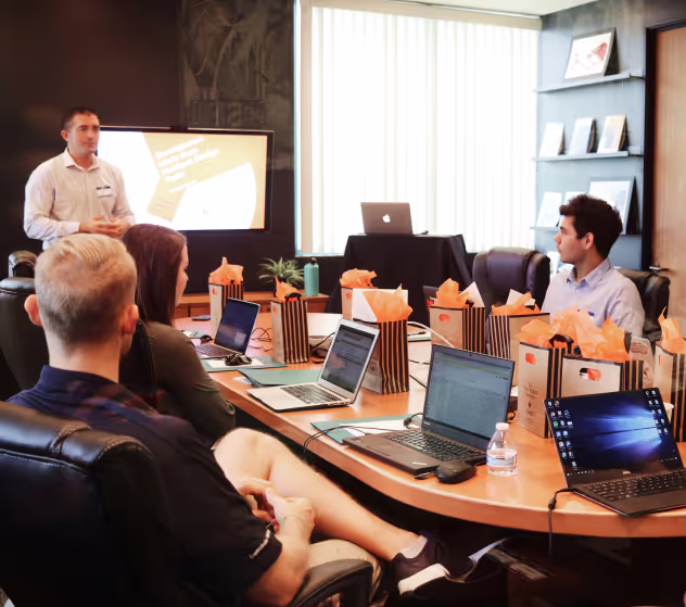 Corporate training or workshop setting in a modern conference room. A presenter stands at the front beside a screen displaying a slide, while four participants seated around a large table engage with laptops and materials. Gift bags with orange tissue paper are arranged in front of each attendee, suggesting a formal workshop or client event. Natural light enters through large vertical blinds behind the presenter.