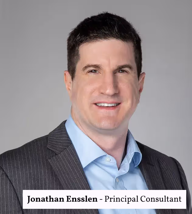 Headshot of Jonathan Ensslen, Principal Consultant. He is wearing a dark suit jacket over a light blue collared shirt, smiling confidently against a neutral gray background. The name and title are displayed in a white text box at the bottom of the image.