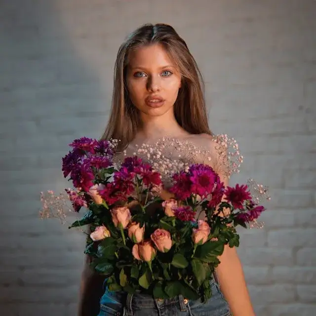 Young woman with long hair standing against a brick wall, wearing a sheer top and jeans, holding a large bouquet of pink and purple flowers in front of her.