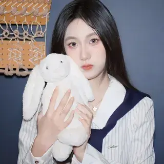 Young woman with long dark hair holding a white stuffed bunny toy in front of a dark blue wall.
