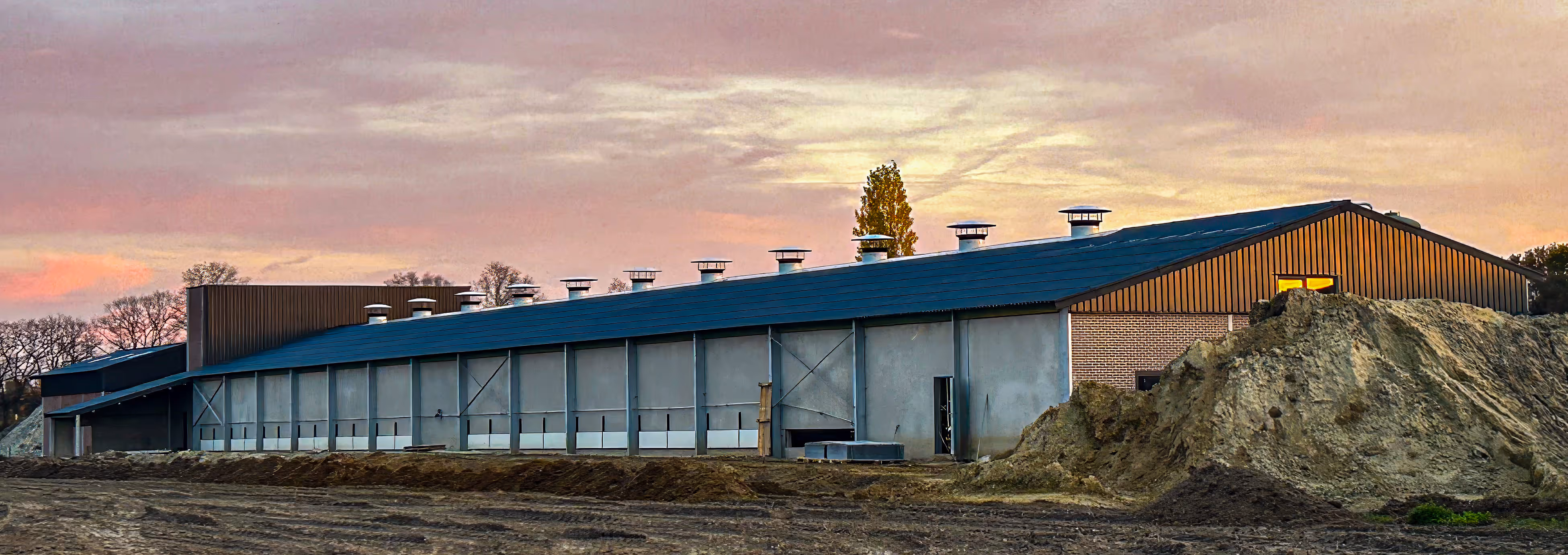 Grote landbouwschuur met ventilatiebuizen op het dak bij zonsondergang.