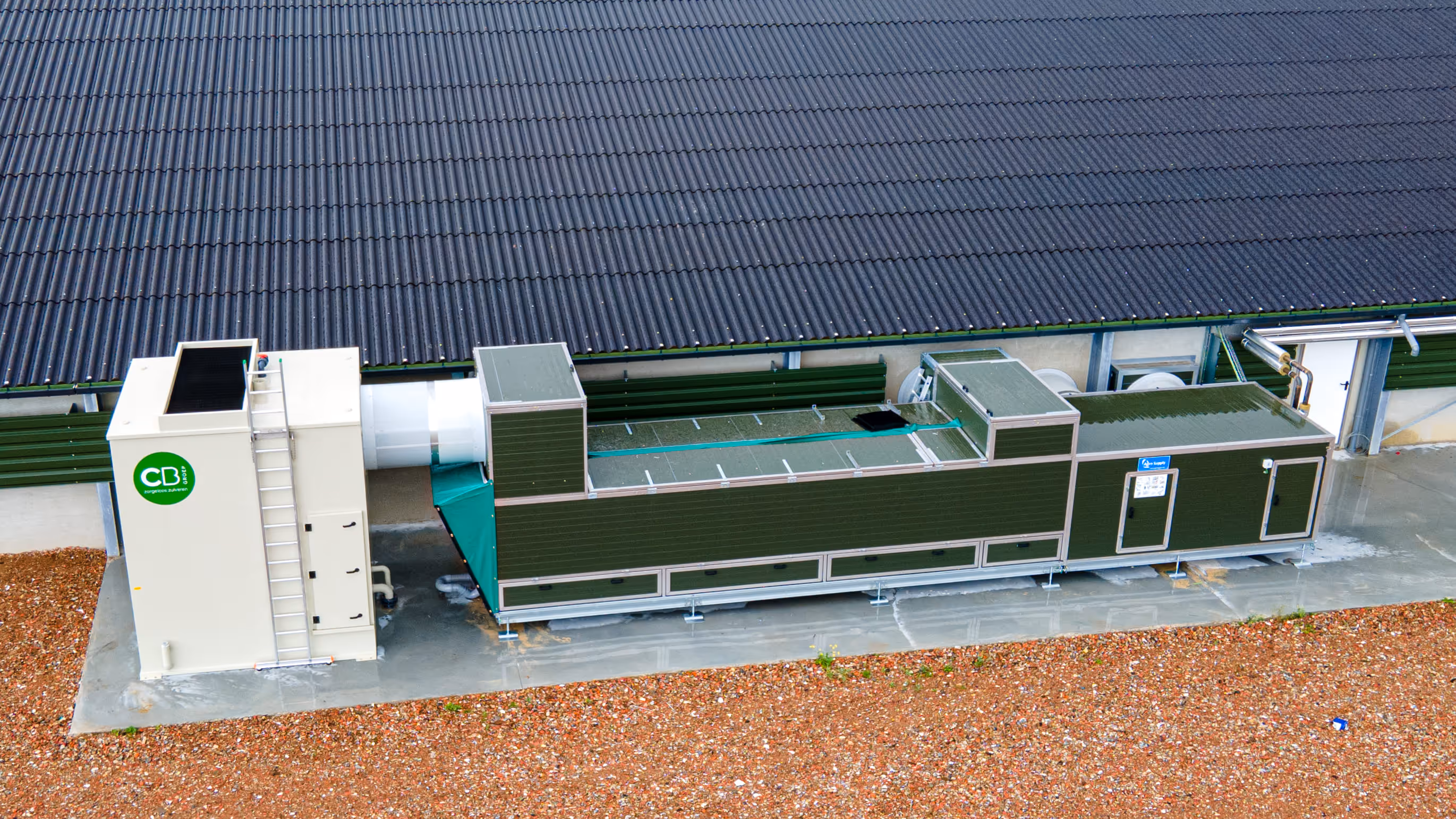 Grote groen-witte industriële HVAC-unit naast een gebouw met een golfplaten dak op een betonnen fundering.